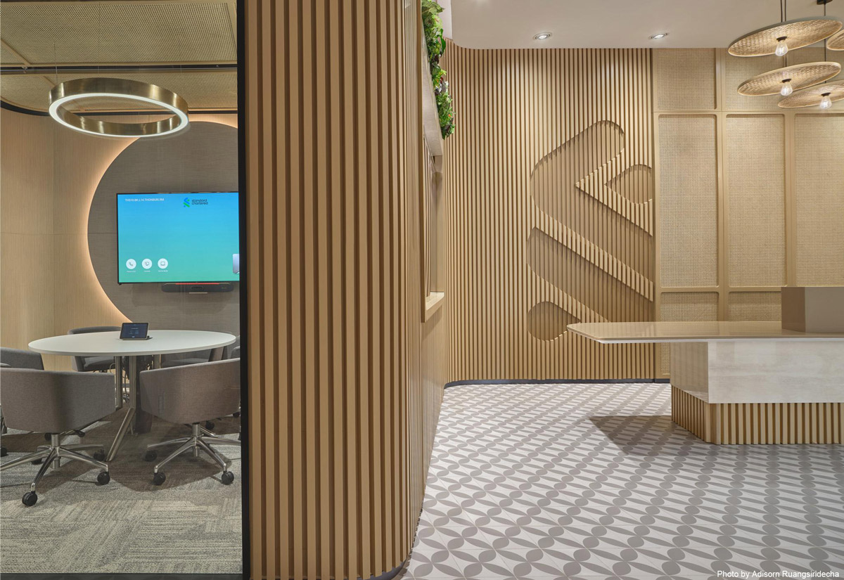 Aedas’s Standard Chartered Bank Bangkok office interior is shown with a human‑centric connect zone comprising timber slat partitions, integrated meeting booth, patterned flooring, and suspended feature lighting. The image highlights modular collaboration spaces, acoustic zoning, and contemporary financial‑workplace interiors designed for flexibility and social interaction.