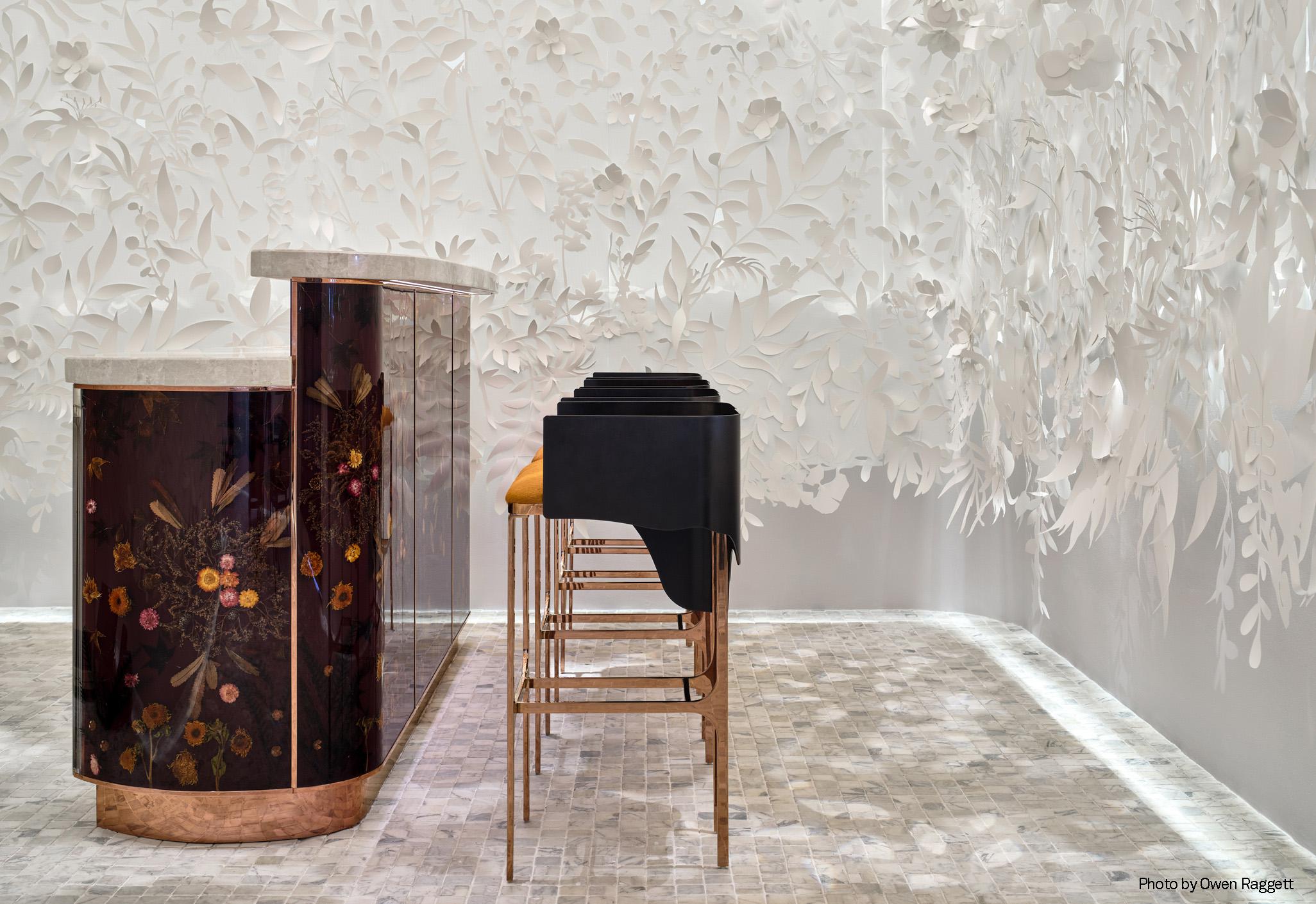 Aedas Interiors’ Yi by Jereme Leung at Raffles Hotel Singapore is shown with an immersive bar and arrival space featuring a sculptural white floral installation, dark lacquered bar counter, refined marble flooring, and minimalist seating. The image highlights an experiential dining interior that blends mythology‑inspired artistry with crafted natural materials to create a layered, contemplative hospitality environment.