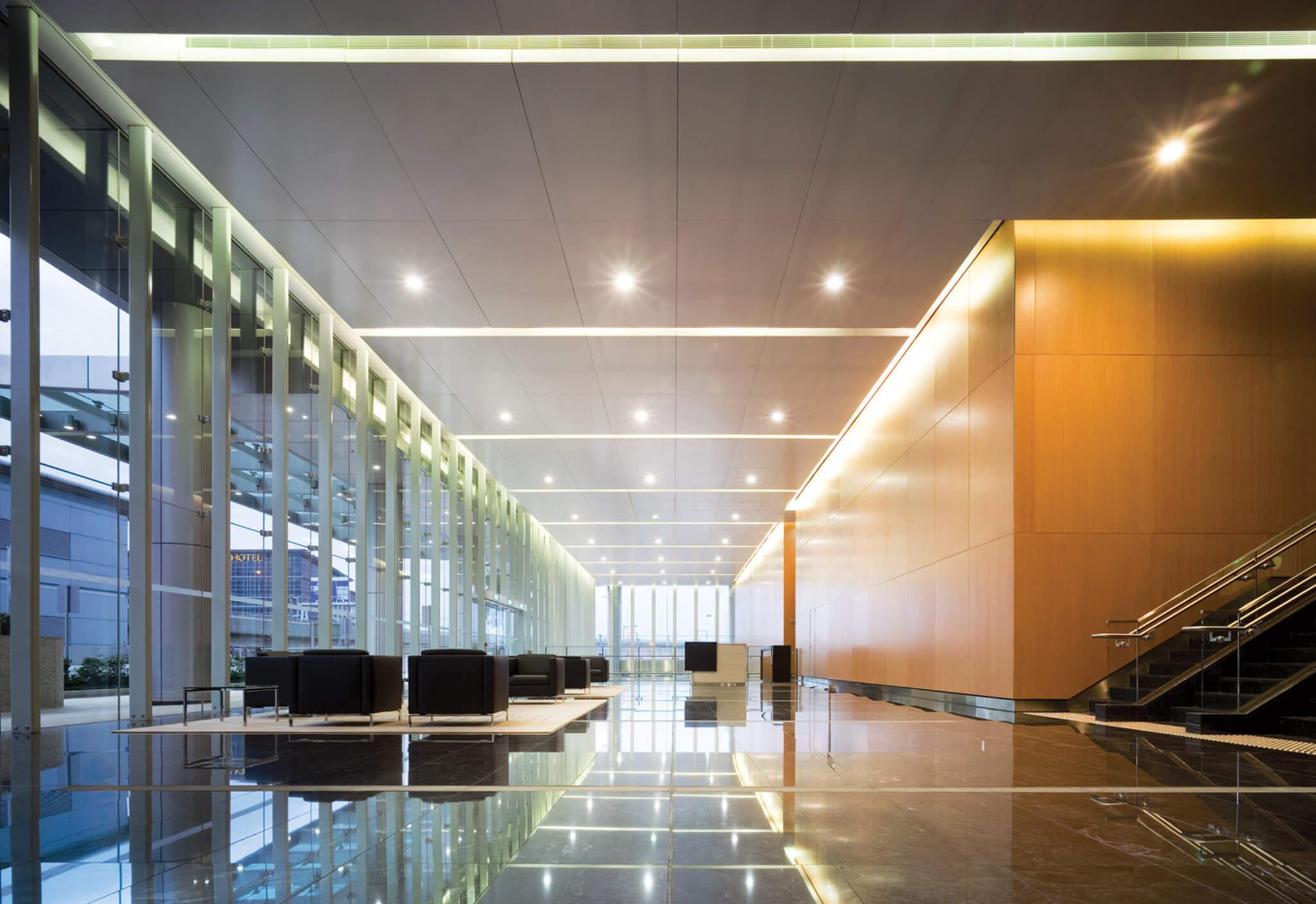 Aedas Interiors’ Airport Authority Hong Kong Headquarters is shown as a contemporary corporate interior with expansive glazed façades, polished stone flooring, and a striking timber‑clad feature wall. The image highlights a light‑filled arrival and circulation space, integrating natural materials with linear ceiling lighting and a calm, spacious layout that reflects an aviation‑linked, modern workplace environment.