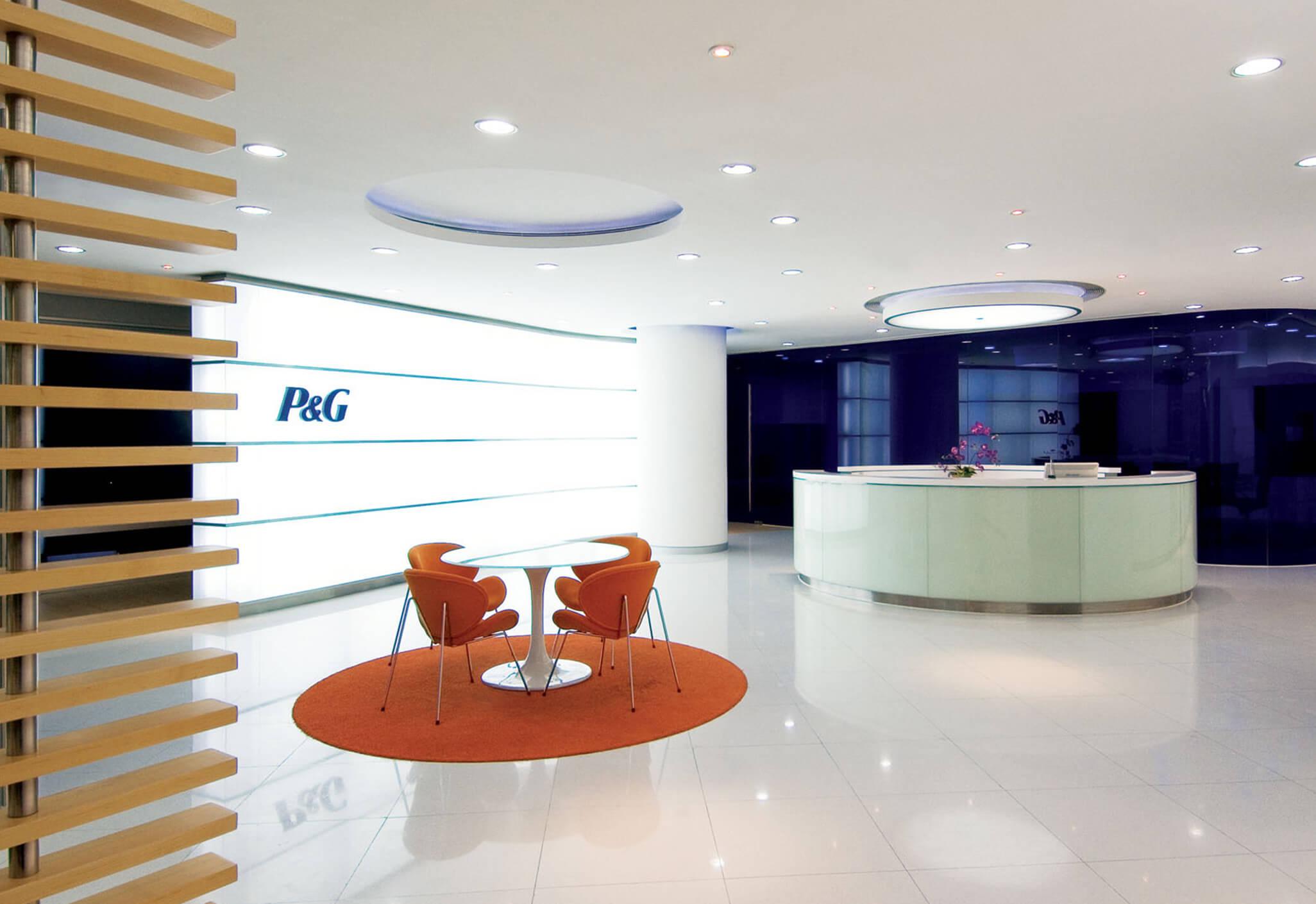 Aedas Interiors’ Procter & Gamble office in Guangzhou is shown with a bright reception and collaboration area featuring a circular white reception desk, curved ceiling elements, vivid accent seating, and brand‑integrated wall graphics. The image highlights a contemporary corporate interior using circular motifs, energised colour palettes, and flexible touchdown spaces to support collaborative work and strong brand identity.