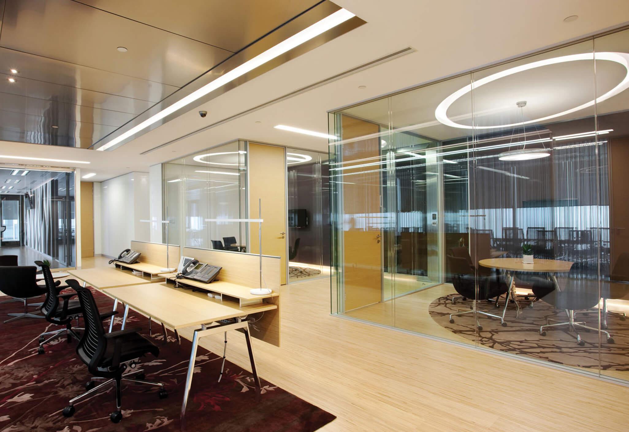 Aedas Interiors’ The Link office at One Landmark East in Hong Kong is shown with a light‑filled collaborative workspace featuring timber flooring, translucent glass meeting rooms, and open workstations. The image highlights an energetic corporate interior with integrated wayfinding, flexible multifunction spaces, and a tree‑pattern feature column reinforcing brand identity within a modern REIT headquarters environment.
