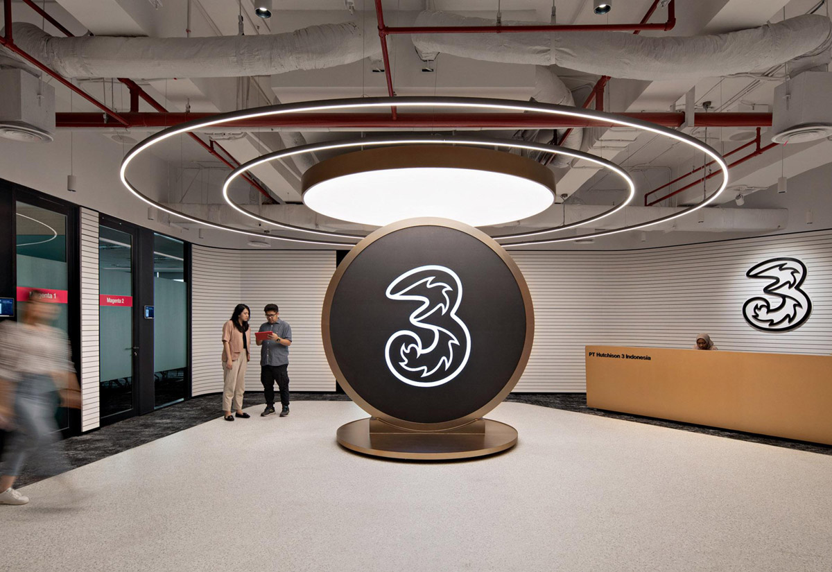 Aedas’s Hutchison 3 Indonesia office in Jakarta is shown with a brand‑centric collaboration zone featuring a sculptural circular centerpiece, halo lighting, curved reception desk, and exposed services ceiling. The image highlights technology‑enabled corporate interiors, event‑ready gathering space, and social workplace design aligned with a telecommunications brand.