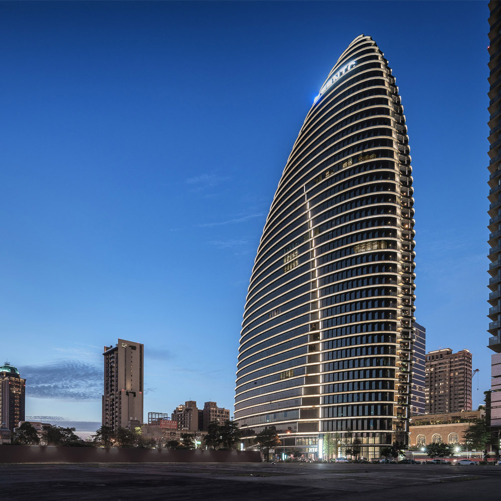 Aedas’ National Trade Center Taichung with oval bamboo‑inspired form and layered horizontal shading façades. Night‑time exterior view of the Aedas‑designed National Trade Center in Taichung, showing an oval high‑rise office tower with horizontally layered floor plates and curved glazing. Continuous horizontal rain covers, fold‑type curtain wall panels, and integrated façade lighting define the silhouette, with vertical green zones and a recessed base connecting the tower to the surrounding urban fabric.