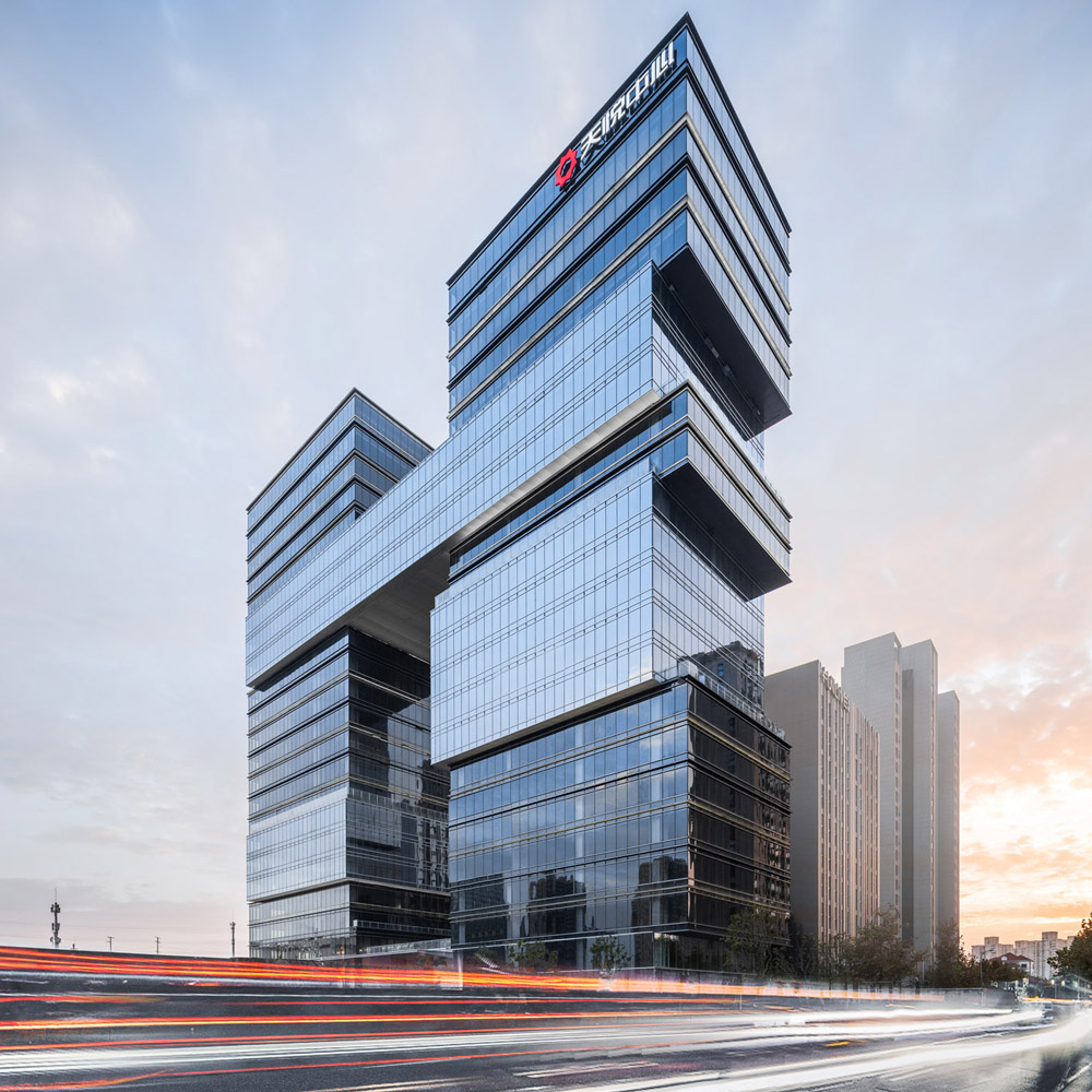 Aedas’ SCE Zhenru office towers with stacked glass volumes, cantilevered terraces and integrated sky connections in Shanghai. Street‑level dusk view of the Aedas‑designed Shanghai SCE Zhenru development, showing two office towers composed of staggered, cantilevered glass volumes. Floor‑to‑ceiling curtain wall façades define stacked blocks of varying sizes, with horizontal projections forming viewing platforms. The towers are visually linked at upper levels and front a primary urban roadway.