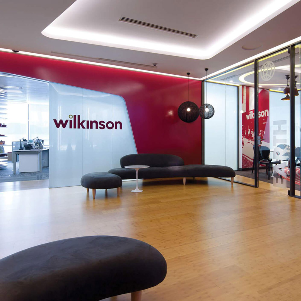 Aedas Interiors’ Wilkinson Asia Headquarters in Hong Kong is shown with a brand‑forward collaborative environment featuring a curved white innovation room, bold red feature walls, timber flooring, and glazed meeting suites. The image highlights flexible lounge seating, strong corporate branding integration, and spatial planning that prioritises informal collaboration and dynamic interaction across the workplace.