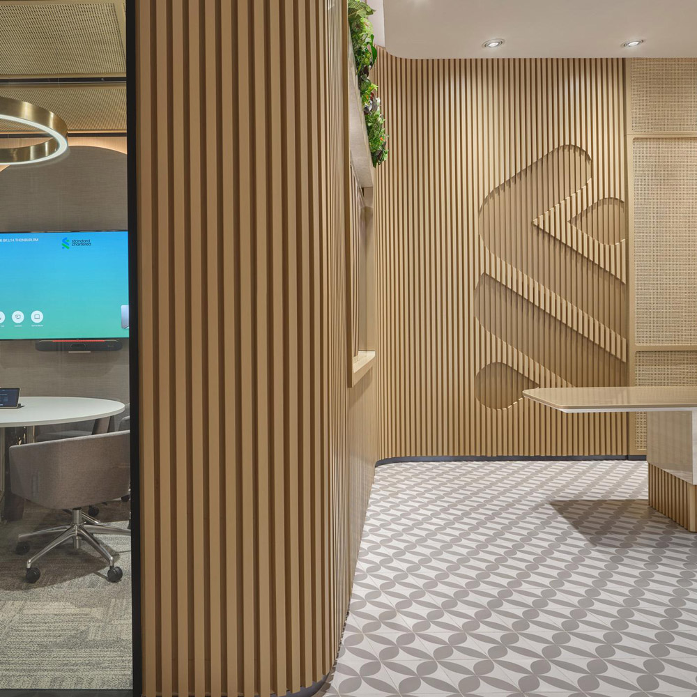 Aedas’s Standard Chartered Bank Bangkok office interior is shown with a human‑centric connect zone comprising timber slat partitions, integrated meeting booth, patterned flooring, and suspended feature lighting. The image highlights modular collaboration spaces, acoustic zoning, and contemporary financial‑workplace interiors designed for flexibility and social interaction.