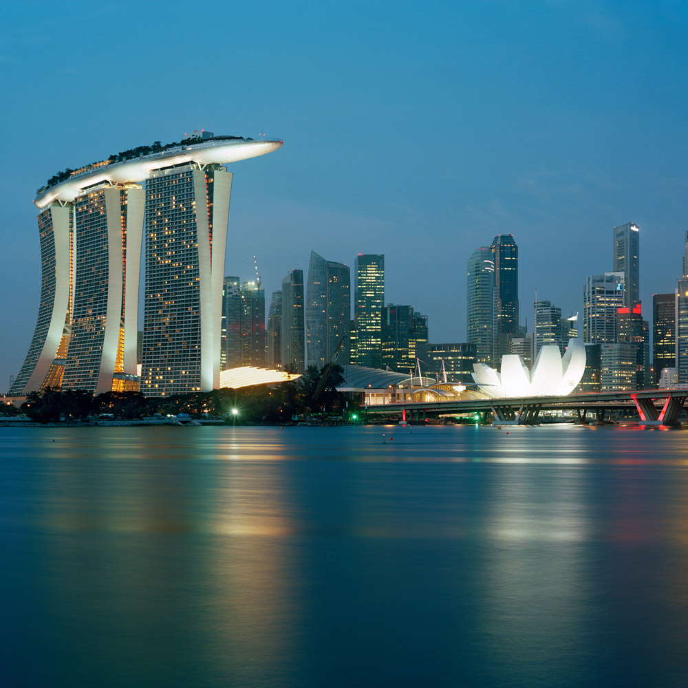 Marina Bay Sands in Singapore is shown as a waterfront integrated resort with three curving hotel towers connected by a dramatic cantilevered rooftop SkyPark. The dusk image highlights the sculptural tower forms, illuminated podium, adjacent ArtScience Museum, and their prominent relationship to Marina Bay and the city skyline.