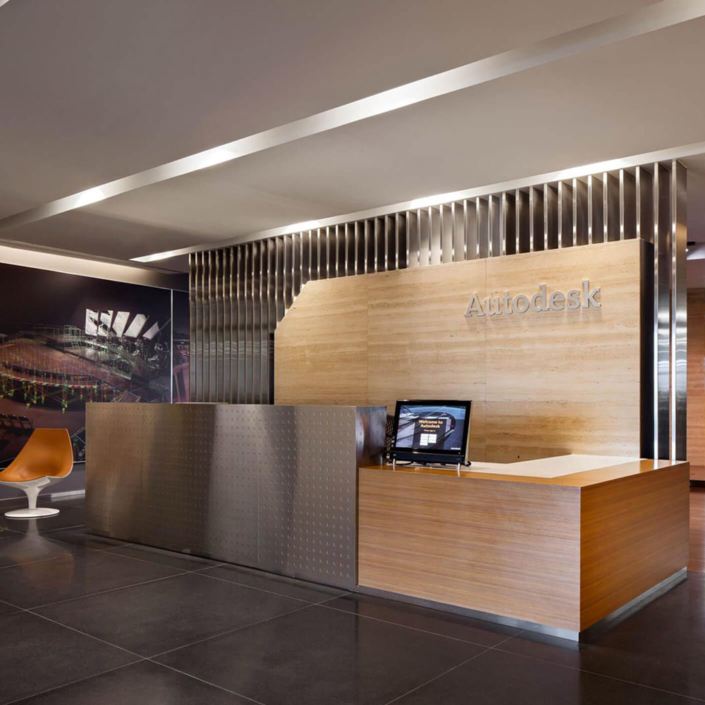 Aedas Interiors’ Autodesk Asia office in Singapore is shown with a refined reception area featuring a timber‑clad counter, perforated metal screen, integrated branding wall, and dark stone flooring. The image highlights a neighbourhood‑inspired workplace interior, combining clean architectural lines, layered materials, and open visual connectivity to support collaboration, client engagement, and a contemporary regional headquarters environment.