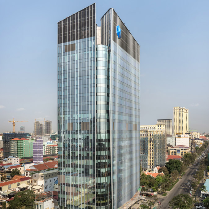 Aedas’ Sathapana Bank office tower with floor‑to‑ceiling glass curtain wall and recessed ground‑floor banking hall. Street‑level view of an Aedas‑designed office tower in central Phnom Penh, showing a rectilinear high‑rise with floor‑to‑ceiling glass curtain wall and a transparent ground‑floor podium. The recessed base accommodates the main banking hall and lobby, defined by structural glazing, landscaped forecourt trees and a shaded pedestrian setback along Preah Norodom Boulevard.