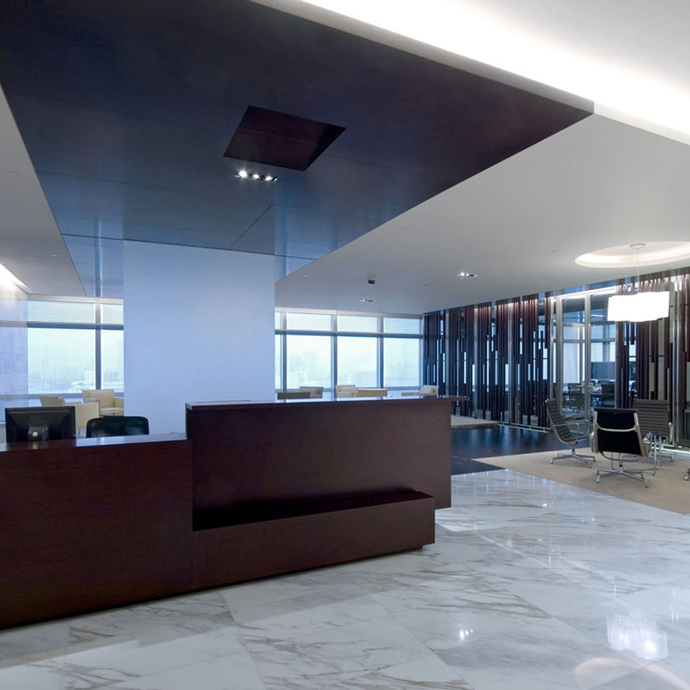 Aedas Interiors’ AIA Asia Headquarters in Hong Kong is shown with an executive reception and lounge environment featuring a dark timber reception counter, polished marble flooring, and semi‑transparent timber screen walls. The image highlights refined corporate interior architecture that blends neutral tones with Asian material influences, creating a calm, prestigious workspace with visual openness and hierarchical zoning across interconnected executive floors.