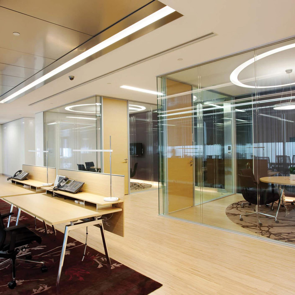 Aedas Interiors’ The Link office at One Landmark East in Hong Kong is shown with a light‑filled collaborative workspace featuring timber flooring, translucent glass meeting rooms, and open workstations. The image highlights an energetic corporate interior with integrated wayfinding, flexible multifunction spaces, and a tree‑pattern feature column reinforcing brand identity within a modern REIT headquarters environment.