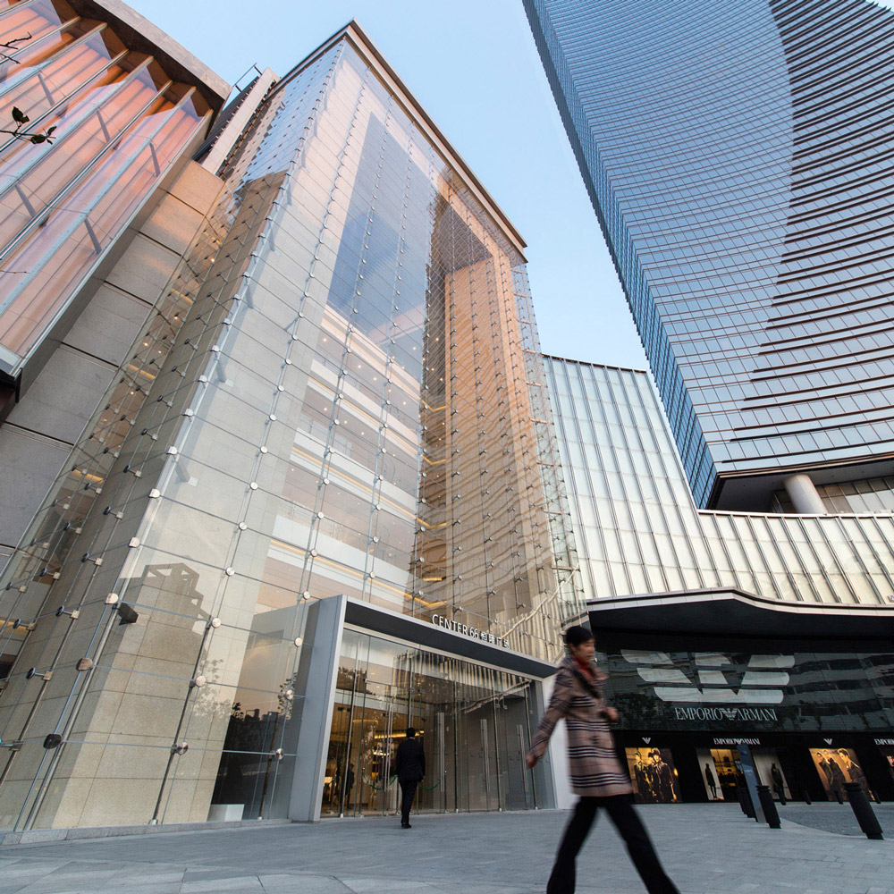 Aedas’s Center 66 Phase 1 in Wuxi is shown as a mixed‑use development with a large glazed retail atrium, horizontally layered mall podium, and a slender high‑rise office tower. The street‑level view highlights transparent façades, active retail frontage, and integration of public space within the city centre.