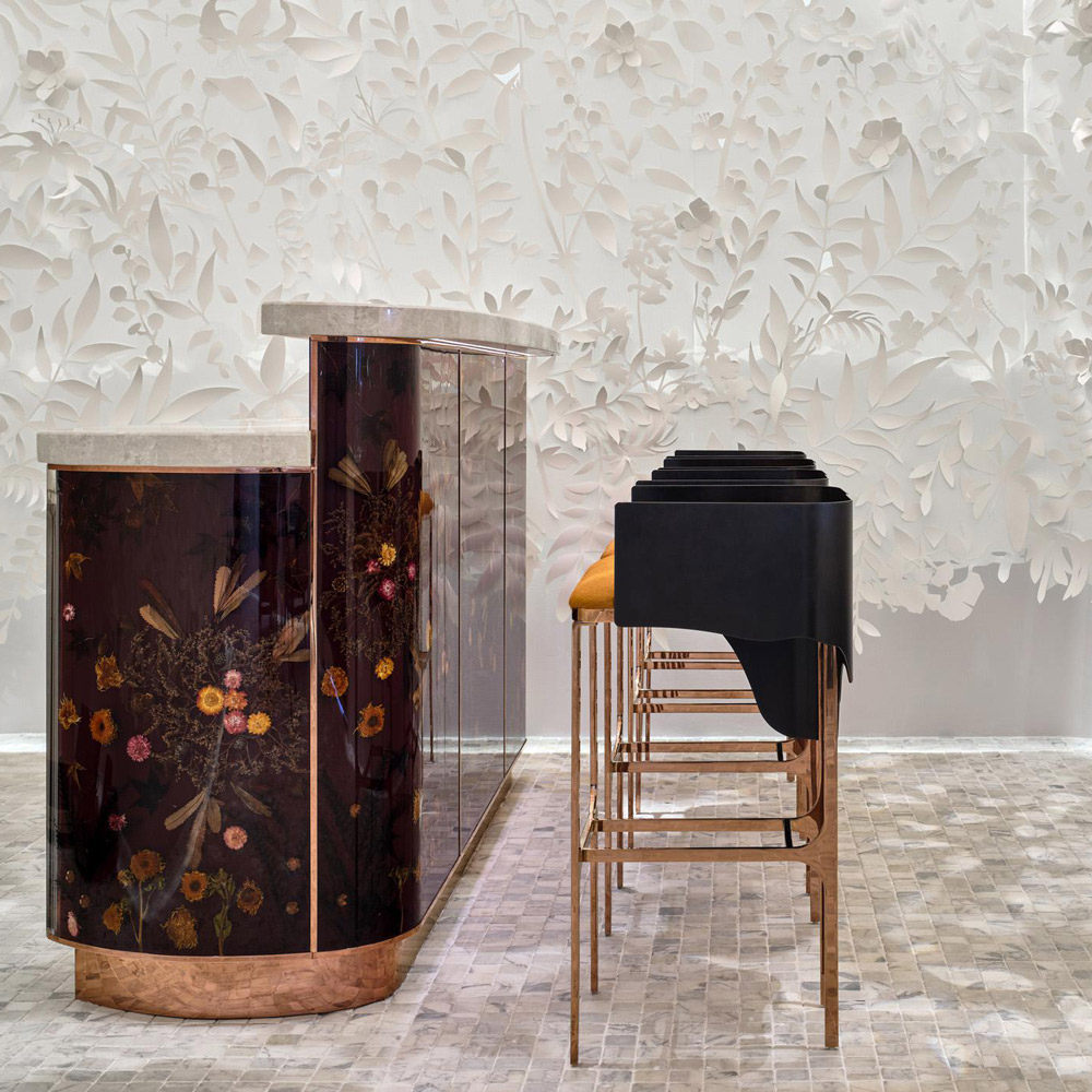 Aedas Interiors’ Yi by Jereme Leung at Raffles Hotel Singapore is shown with an immersive bar and arrival space featuring a sculptural white floral installation, dark lacquered bar counter, refined marble flooring, and minimalist seating. The image highlights an experiential dining interior that blends mythology‑inspired artistry with crafted natural materials to create a layered, contemplative hospitality environment.