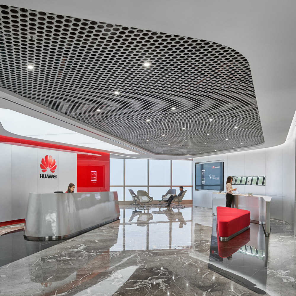 Aedas Interiors’ Huawei Hanoi office is shown with a refined reception environment featuring sculpted metallic counters, a perforated geometric ceiling panel, polished stone flooring, and integrated brand graphics. The image highlights a cohesive corporate interior language enriched with local material accents, varied spatial identities, and colour infusions that create a welcoming, contemporary workplace aligned with Huawei’s global design ethos.