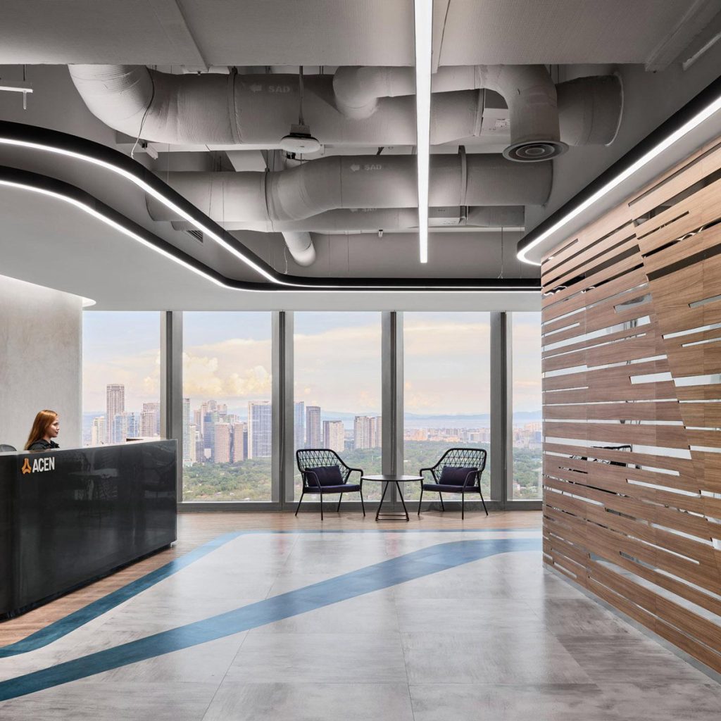 Aedas’s ACEN office in Manila is shown with a contemporary reception and social interior featuring a curved reception counter, vertically layered timber wall, fluid LED ceiling lighting, exposed services, and floor graphics suggesting energy flow. The image highlights sustainability‑driven corporate interior architecture and adaptable workplace design.