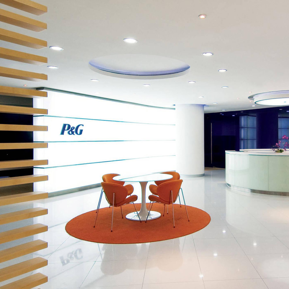 Aedas Interiors’ Procter & Gamble office in Guangzhou is shown with a bright reception and collaboration area featuring a circular white reception desk, curved ceiling elements, vivid accent seating, and brand‑integrated wall graphics. The image highlights a contemporary corporate interior using circular motifs, energised colour palettes, and flexible touchdown spaces to support collaborative work and strong brand identity.