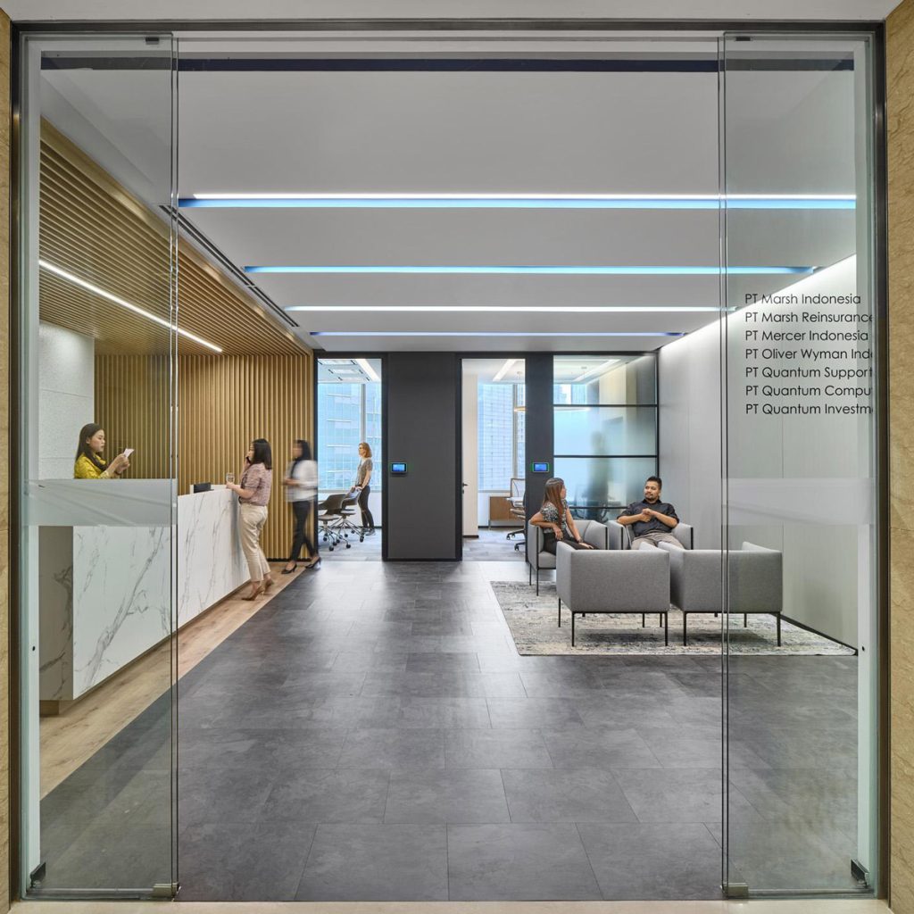 Aedas Interiors’ Marsh & McLennan Jakarta Smart Office is shown as a contemporary, glass‑fronted staff hub with integrated reception, lounge seating, and open circulation. The image highlights an agile, unassigned workplace interior combining neutral tones, timber accents, and local materials, designed to support connectivity, informal collaboration, and diverse work settings within a vibrant, technology‑enabled corporate environment.