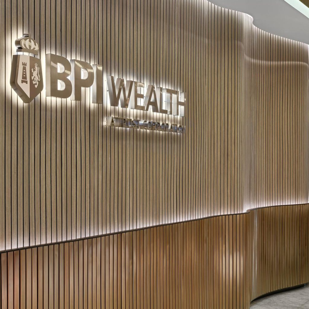 Aedas’s Bank of The Philippine Islands interior in Manila is shown with a sculpted reception area featuring vertically ribbed timber wall panels, a layered curved counter, and integrated illuminated BPI Wealth signage. The image highlights refined material detailing, corporate brand expression, acoustic wall treatment, and a contemporary banking workplace interior.