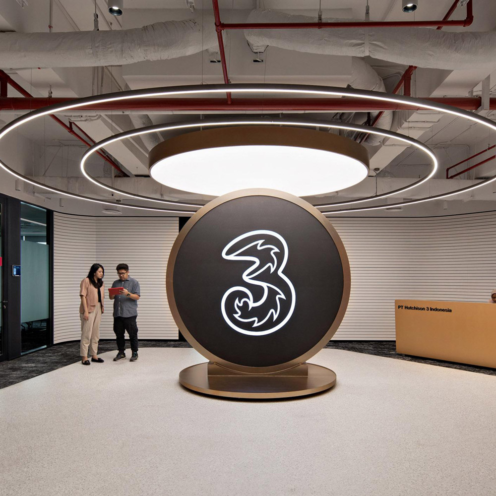 Aedas’s Hutchison 3 Indonesia office in Jakarta is shown with a brand‑centric collaboration zone featuring a sculptural circular centerpiece, halo lighting, curved reception desk, and exposed services ceiling. The image highlights technology‑enabled corporate interiors, event‑ready gathering space, and social workplace design aligned with a telecommunications brand.
