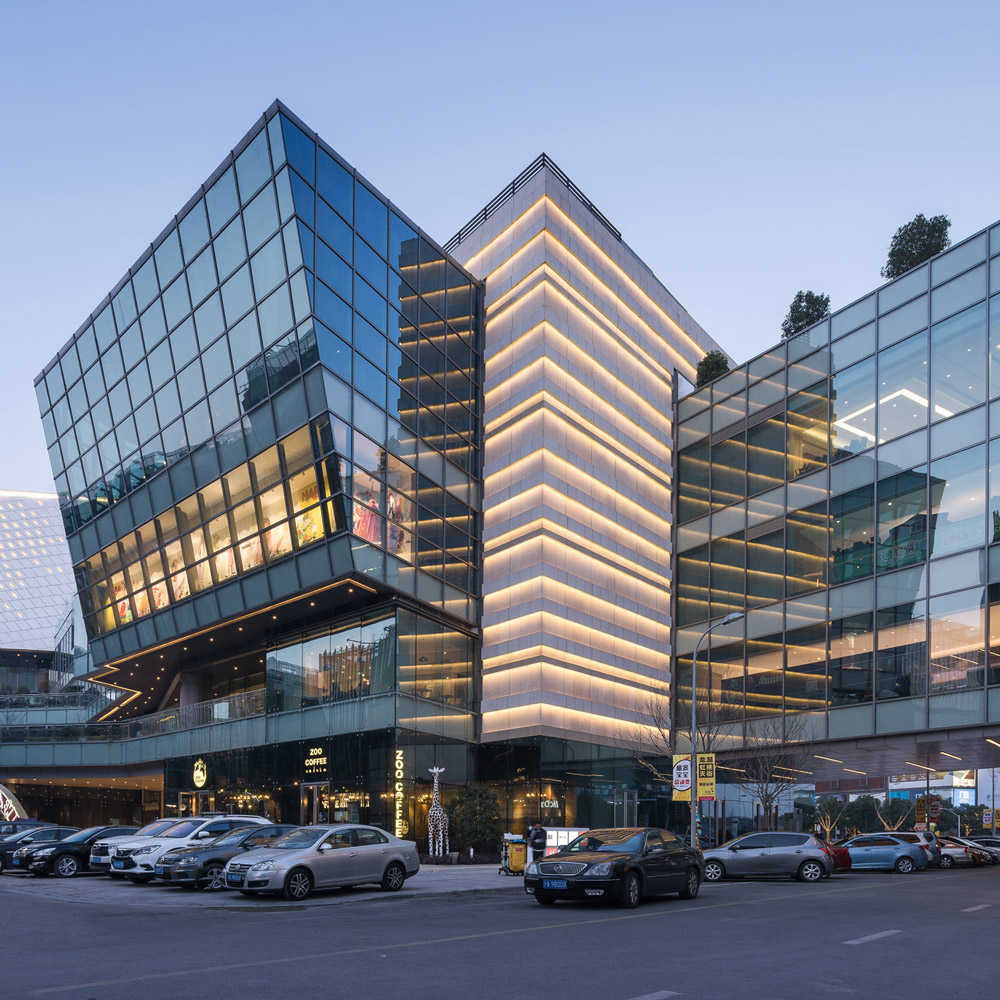 Aedas’ Longfor Hongqiao mixed‑use complex with folded glass façades linking retail podium, office volumes and hotel tower in CBD. Street‑level view of an Aedas‑designed mixed‑use development showing interlocking retail, office and hotel volumes. The podium adopts a folded, linear form with extensive floor‑to‑ceiling glazing and continuous horizontal slab edges. Upper blocks rise from the base with articulated glass façades, creating layered public terraces and exposed circulation zones facing a central business district street.