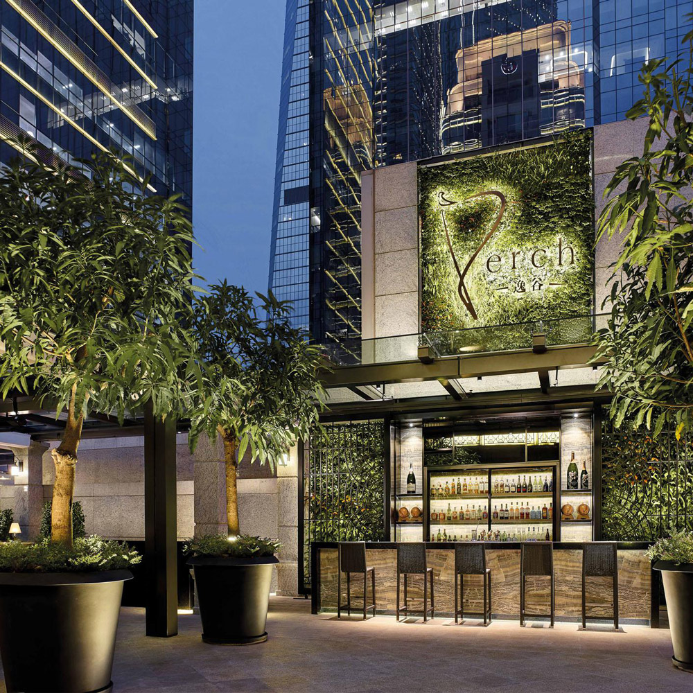 Aedas Interiors’ Perch at The Langham Shenzhen is shown as an elevated outdoor bar with lush potted trees, a green living wall façade, and a central illuminated bar pavilion. The image highlights a refined hospitality environment that blends landscape elements, warm lighting, and contemporary stone detailing, creating an intimate urban terrace setting framed by surrounding high‑rise towers.