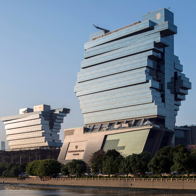 Aedas’ Pazhou exhibition complex with stacked cantilevered volumes and glass‑clad mixed‑use towers in Guangzhou. Riverside view of an Aedas‑designed exhibition and mixed‑use complex on Pazhou Island, Guangzhou, showing two large buildings composed of stacked elongated volumes. The façades combine extensive glass curtain walls with solid cladding panels. One structure supports a high‑rise hotel tower above exhibition halls, while the other integrates offices over a commercial exhibition base.