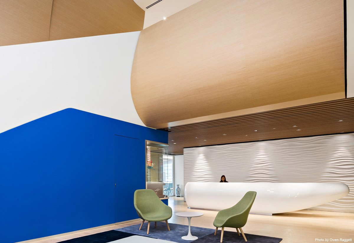 Aedas Interiors’ DNV GL Technology Centre in Singapore is shown with an open reception and atrium space featuring a sculptural curved timber ceiling, wave‑textured white feature wall, and container‑inspired reception counter. The image highlights organic, maritime‑influenced interior architecture that connects floors visually, supports social interaction, and reflects a sustainable, innovation‑driven workplace environment.