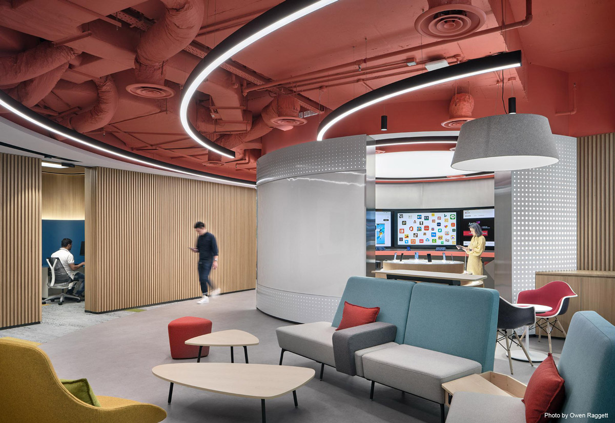 Aedas Interiors’ DIGIX Lab in Singapore is shown as an experiential technology showroom with a bold coral‑painted exposed ceiling, curved LED lighting, modular seating, and interactive app display zones. The image highlights flexible interior planning, strong brand colour integration, and a hybrid environment supporting collaboration, demonstration, and training functions.