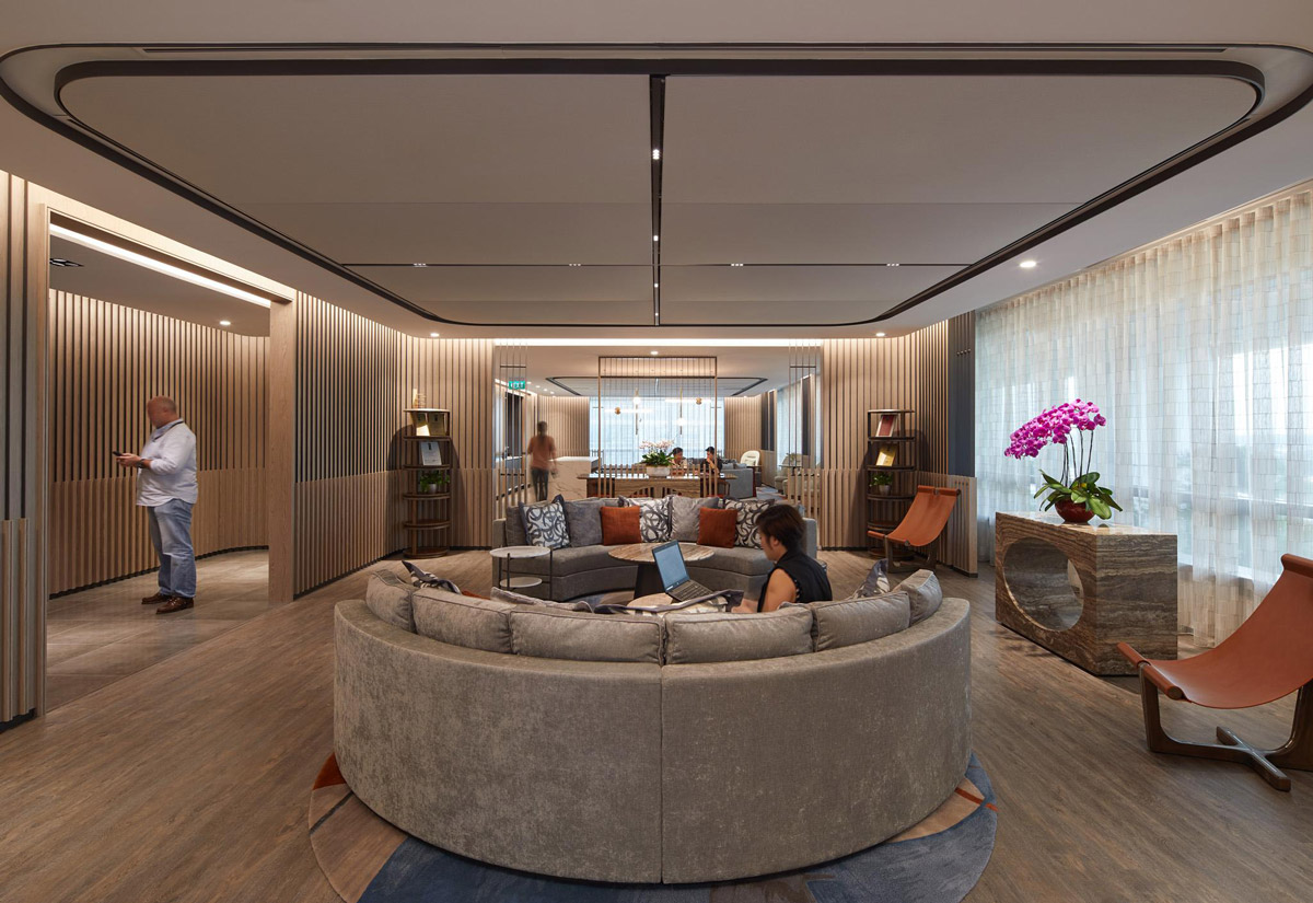 Aedas Interiors’ Singapore Office is shown with a central collaborative lounge featuring circular modular seating, vertical timber slat partitions, integrated shelving, and layered ceiling lighting. The image highlights a contemporary workplace interior designed to foster peer collaboration, informal meetings, and flexible work modes within a large design studio environment.