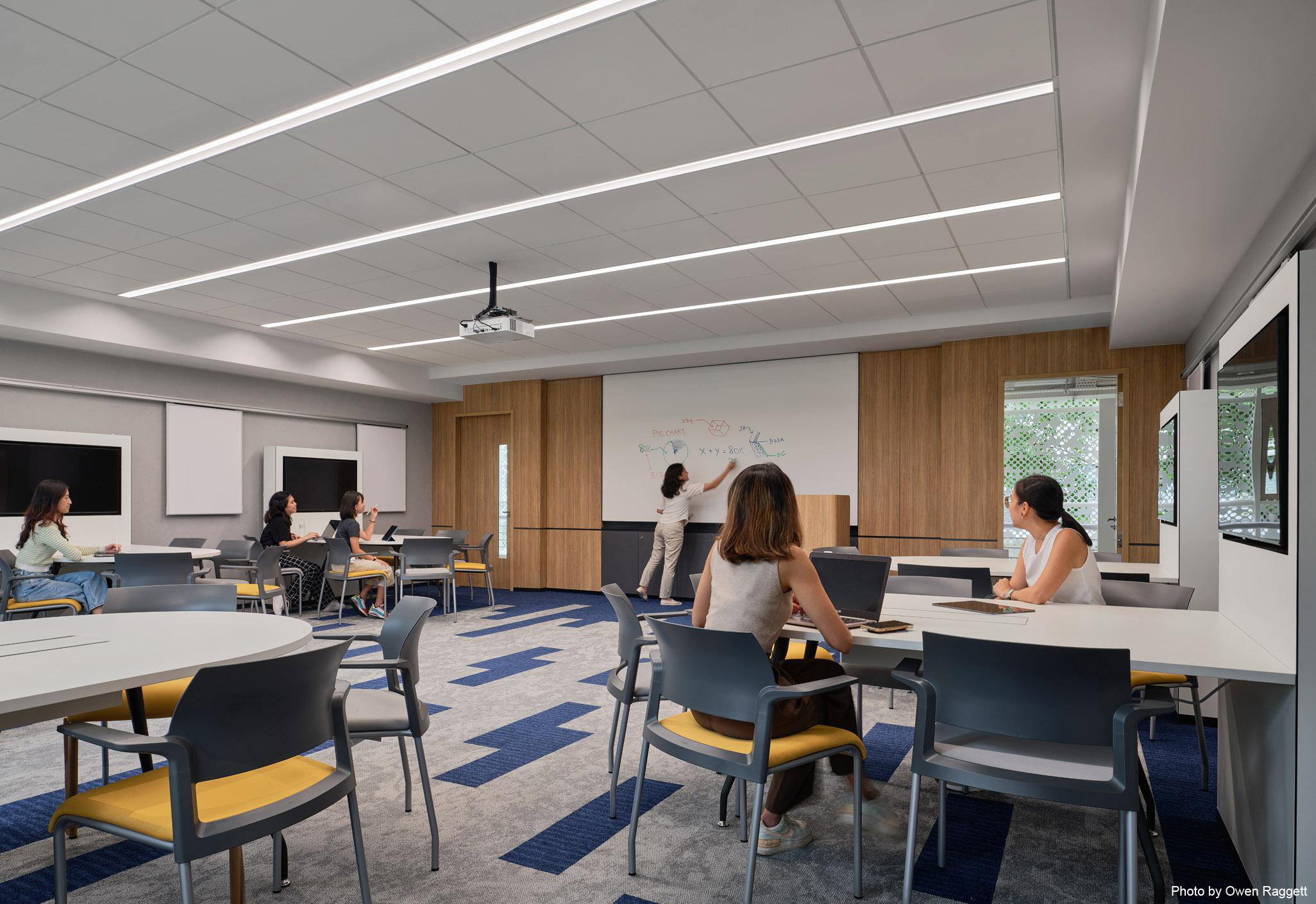 Aedas’s James Cook University Singapore interior is shown as a contemporary flexible teaching classroom with reconfigurable tables, integrated audiovisual systems, acoustic ceiling panels, and perimeter glazing. The image highlights technology‑enabled learning spaces, zoned circulation, and interior architecture designed to support both collaborative and focused study modes.