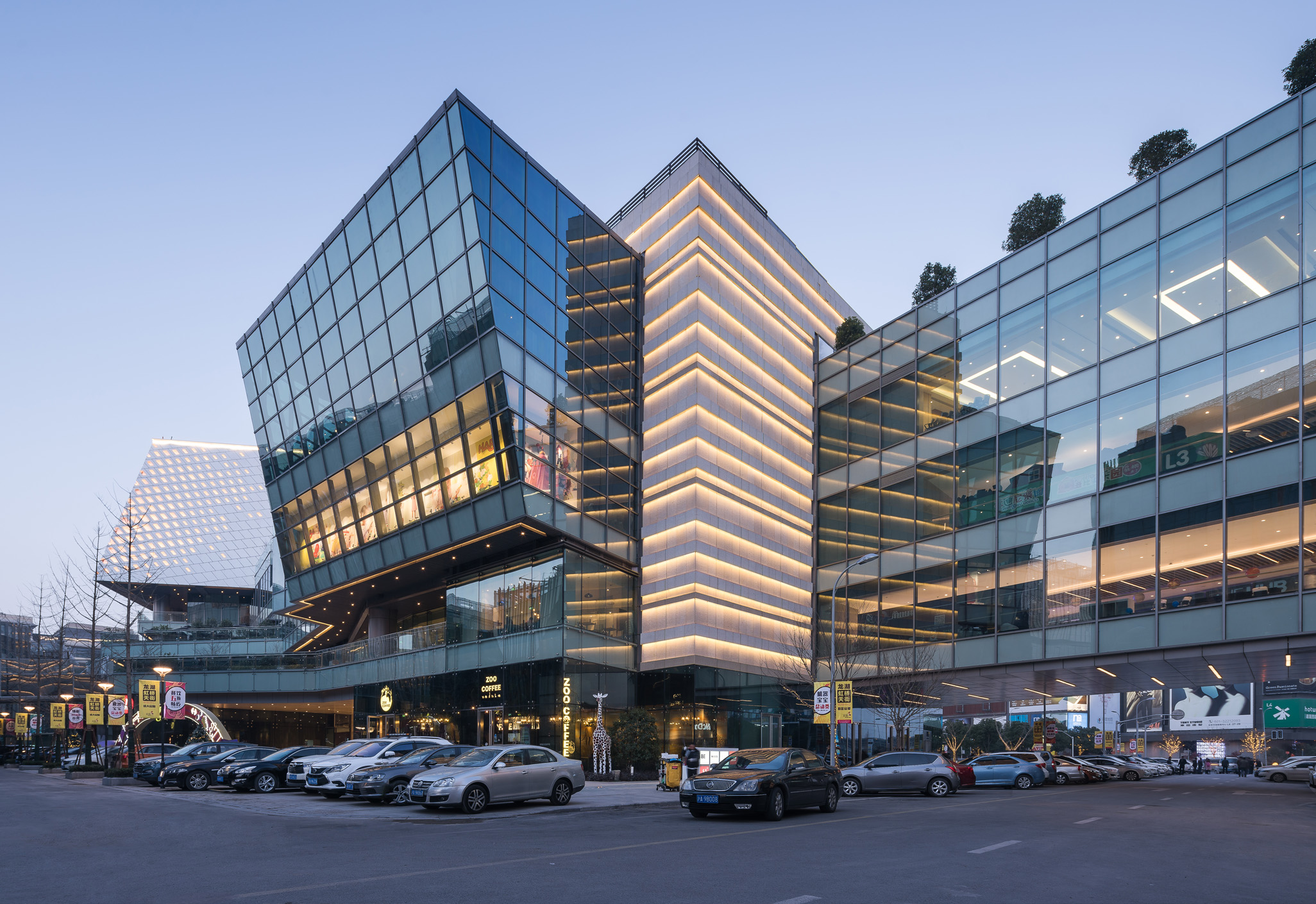 Aedas’ Longfor Hongqiao mixed‑use complex with folded glass façades linking retail podium, office volumes and hotel tower in CBD. Street‑level view of an Aedas‑designed mixed‑use development showing interlocking retail, office and hotel volumes. The podium adopts a folded, linear form with extensive floor‑to‑ceiling glazing and continuous horizontal slab edges. Upper blocks rise from the base with articulated glass façades, creating layered public terraces and exposed circulation zones facing a central business district street.