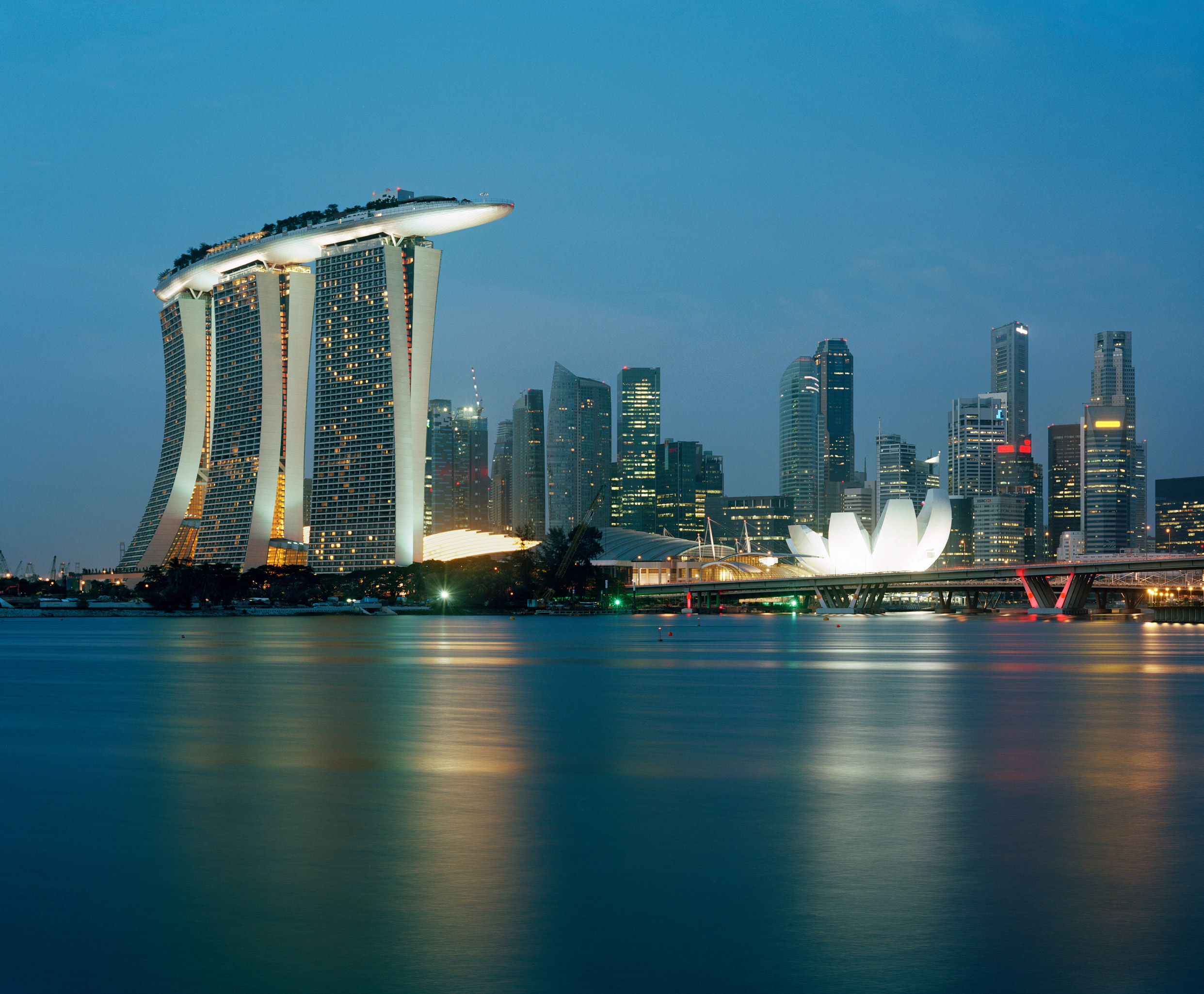Marina Bay Sands in Singapore is shown as a waterfront integrated resort with three curving hotel towers connected by a dramatic cantilevered rooftop SkyPark. The dusk image highlights the sculptural tower forms, illuminated podium, adjacent ArtScience Museum, and their prominent relationship to Marina Bay and the city skyline.