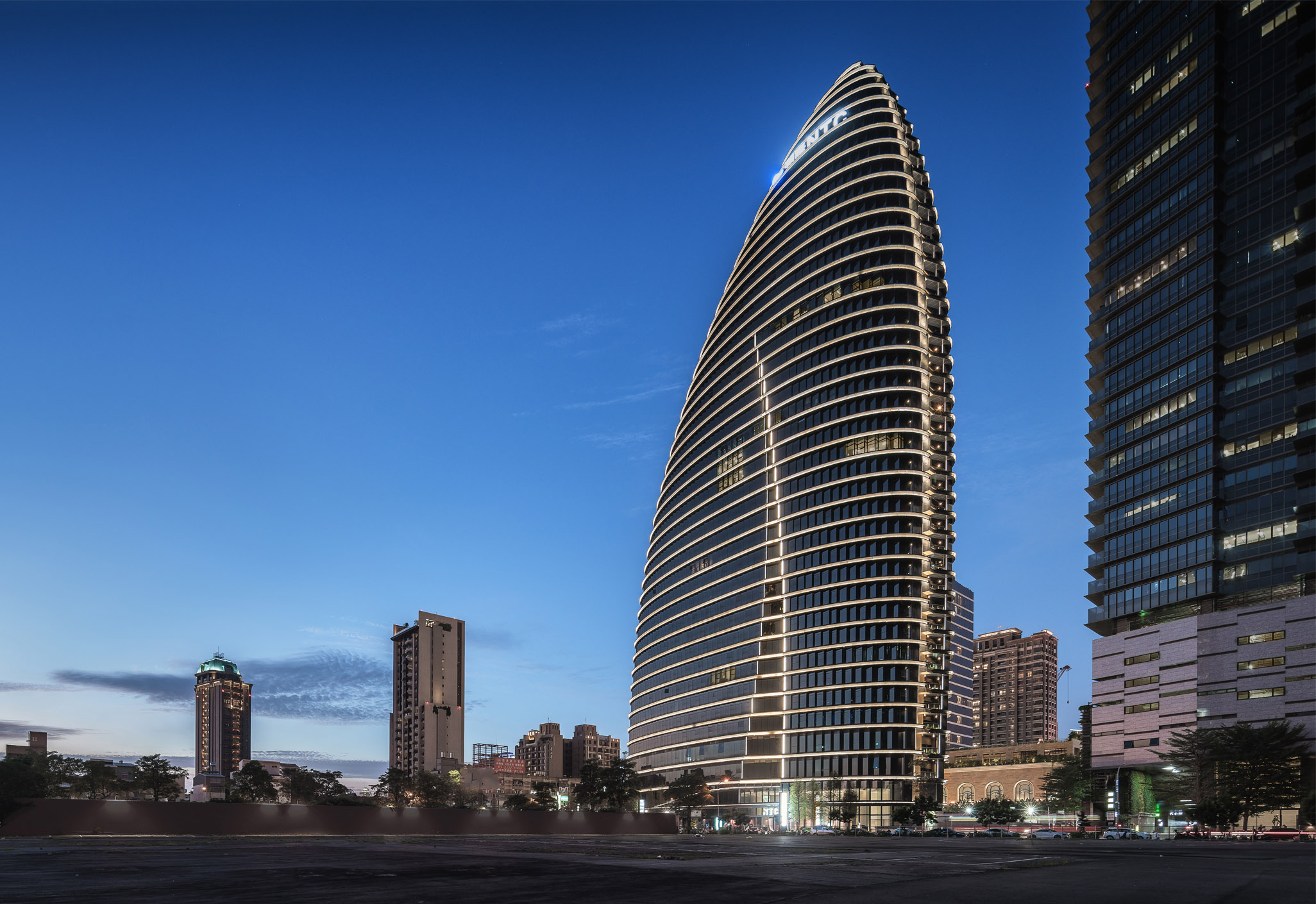 Aedas’ National Trade Center Taichung with oval bamboo‑inspired form and layered horizontal shading façades. Night‑time exterior view of the Aedas‑designed National Trade Center in Taichung, showing an oval high‑rise office tower with horizontally layered floor plates and curved glazing. Continuous horizontal rain covers, fold‑type curtain wall panels, and integrated façade lighting define the silhouette, with vertical green zones and a recessed base connecting the tower to the surrounding urban fabric.