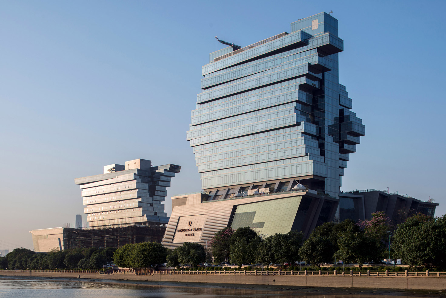 Aedas’ Pazhou exhibition complex with stacked cantilevered volumes and glass‑clad mixed‑use towers in Guangzhou. Riverside view of an Aedas‑designed exhibition and mixed‑use complex on Pazhou Island, Guangzhou, showing two large buildings composed of stacked elongated volumes. The façades combine extensive glass curtain walls with solid cladding panels. One structure supports a high‑rise hotel tower above exhibition halls, while the other integrates offices over a commercial exhibition base.