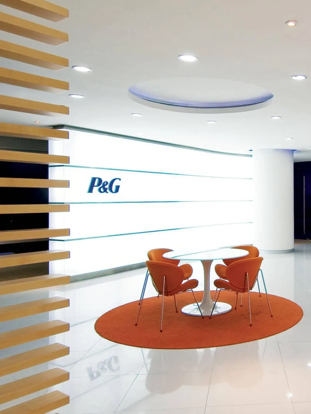 Aedas Interiors’ Procter & Gamble office in Guangzhou is shown with a bright reception and collaboration area featuring a circular white reception desk, curved ceiling elements, vivid accent seating, and brand‑integrated wall graphics. The image highlights a contemporary corporate interior using circular motifs, energised colour palettes, and flexible touchdown spaces to support collaborative work and strong brand identity.