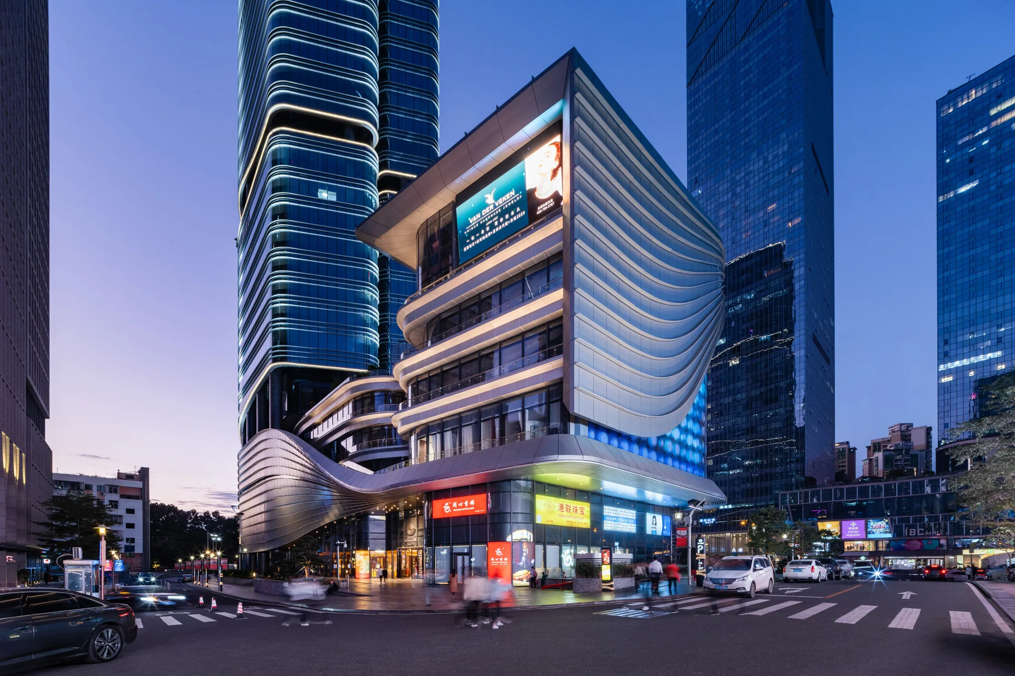 Aedas’ SHUIBEI International Center with bamboo‑inspired tower massing rising above a multi‑level retail podium in Shenzhen. Evening street‑level view of the Aedas‑designed SHUIBEI International Center in Shenzhen, showing a super‑high‑rise office tower with vertically ascending, curved glass façades. The tower rises from a sculpted podium with sweeping horizontal bands, integrated lighting, and active retail frontages, forming a mixed‑use commercial landmark within a dense urban regeneration district.