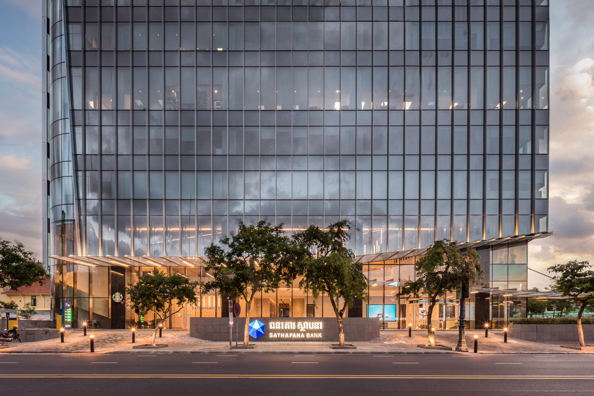 Aedas’ Sathapana Bank office tower with floor‑to‑ceiling glass curtain wall and recessed ground‑floor banking hall. Street‑level view of an Aedas‑designed office tower in central Phnom Penh, showing a rectilinear high‑rise with floor‑to‑ceiling glass curtain wall and a transparent ground‑floor podium. The recessed base accommodates the main banking hall and lobby, defined by structural glazing, landscaped forecourt trees and a shaded pedestrian setback along Preah Norodom Boulevard.