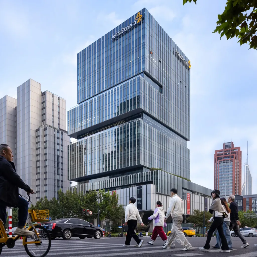 Aedas’ Shanghai EDGE office tower with stepped glass façade and cantilevered retail volume above a public plaza. Street‑level view of Aedas‑designed Shanghai EDGE, showing a vertically stacked office tower with floor‑to‑ceiling glass curtain walls and expressed horizontal setbacks forming external terraces. A large cantilevered retail volume projects over a public plaza at ground level, directly adjacent to a metro entrance, with pedestrian crossings and urban traffic integrated into the streetscape.