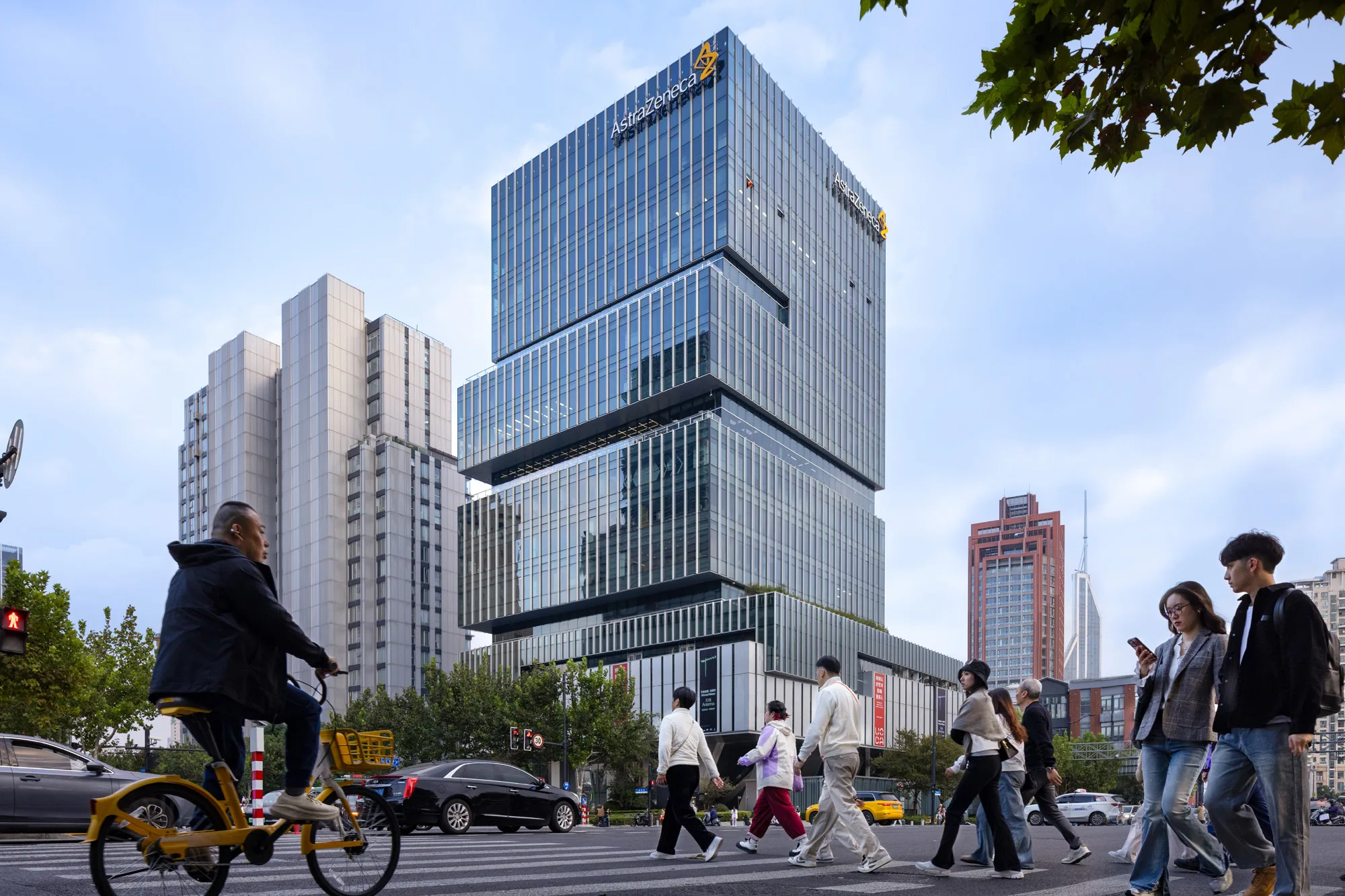 Aedas’ Shanghai EDGE office tower with stepped glass façade and cantilevered retail volume above a public plaza. Street‑level view of Aedas‑designed Shanghai EDGE, showing a vertically stacked office tower with floor‑to‑ceiling glass curtain walls and expressed horizontal setbacks forming external terraces. A large cantilevered retail volume projects over a public plaza at ground level, directly adjacent to a metro entrance, with pedestrian crossings and urban traffic integrated into the streetscape.