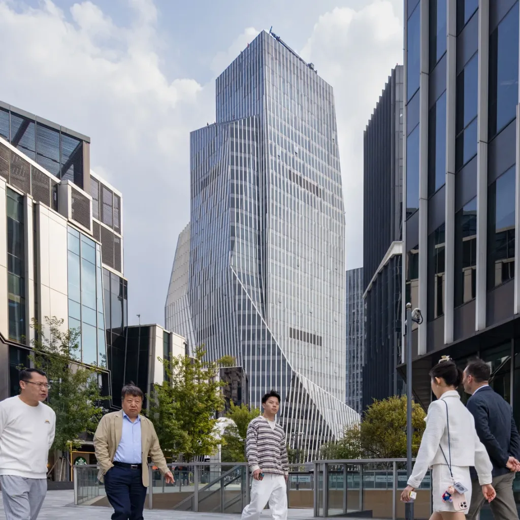 Aedas’ IM Shanghai cultural complex with sculpted glass towers, void‑solid façades and layered urban plaza in Changning. Street‑level view of Aedas‑designed IM Shanghai in Changning District, showing a composition of glass‑clad office towers, angular cultural volumes and low‑rise pavilions. The façades alternate between transparent glazing and solid cladding, forming layered massing around an elevated public plaza with bridges, terraces and pedestrian circulation connecting cultural, office and retail elements.