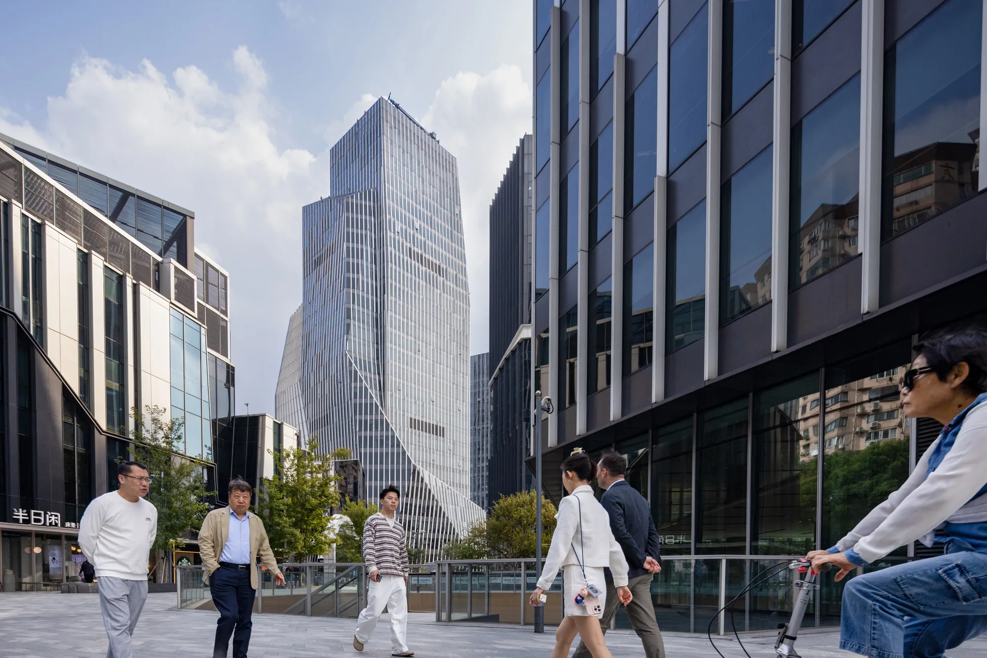 Aedas’ IM Shanghai cultural complex with sculpted glass towers, void‑solid façades and layered urban plaza in Changning. Street‑level view of Aedas‑designed IM Shanghai in Changning District, showing a composition of glass‑clad office towers, angular cultural volumes and low‑rise pavilions. The façades alternate between transparent glazing and solid cladding, forming layered massing around an elevated public plaza with bridges, terraces and pedestrian circulation connecting cultural, office and retail elements.