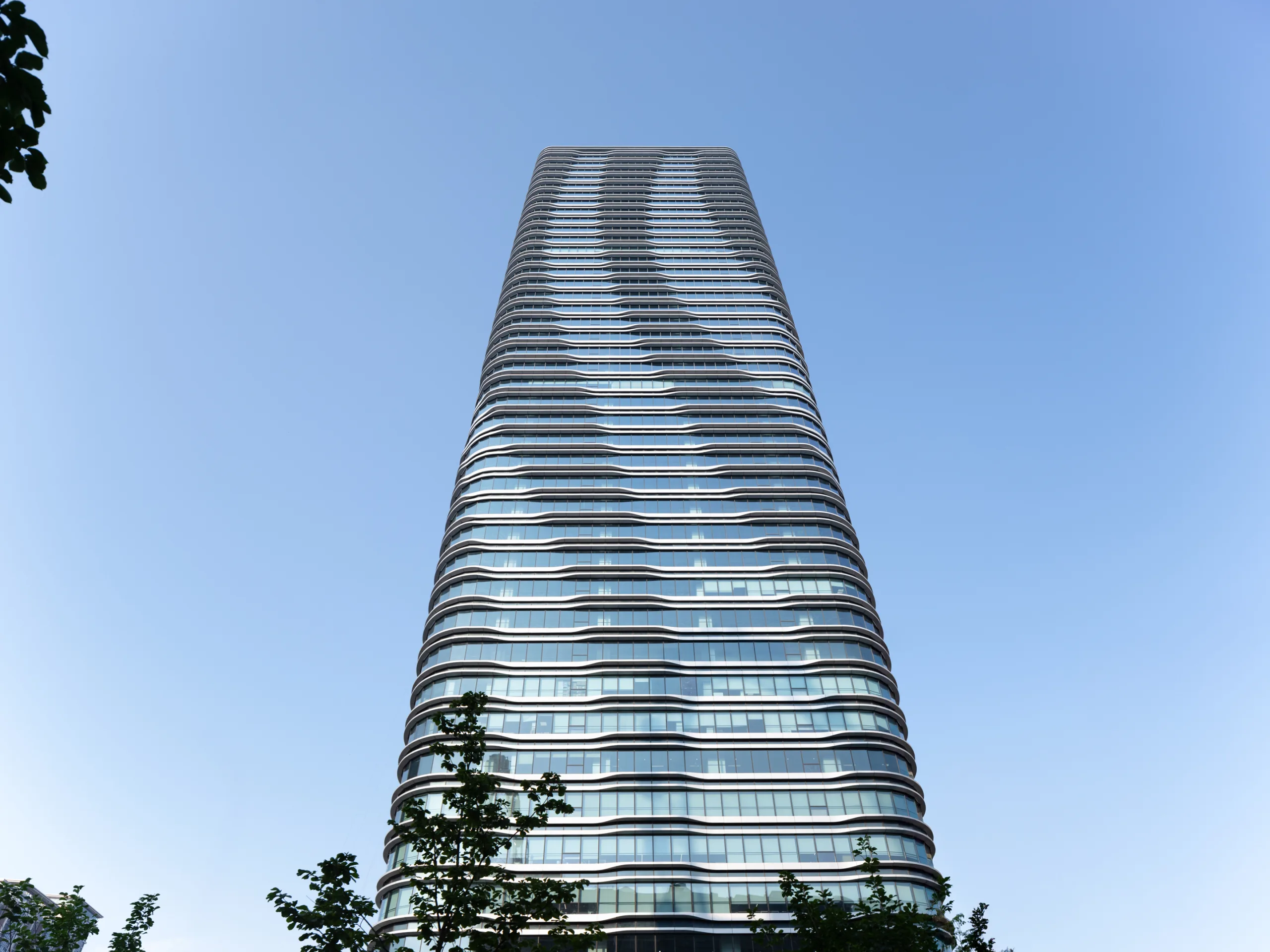 Aedas’ COFCO Joy City Phase 2 office tower with layered glass façade rising above adaptive‑reuse campus in Shanghai. Low‑angle exterior view of Aedas‑designed Shanghai COFCO Joy City Phase 2, showing a tall Grade‑A office tower articulated by floor‑to‑ceiling glass curtain walls and undulating horizontal façade bands. The tower is integrated with repurposed low‑rise buildings at podium level, forming a cohesive mixed‑use headquarters campus within an urban district.