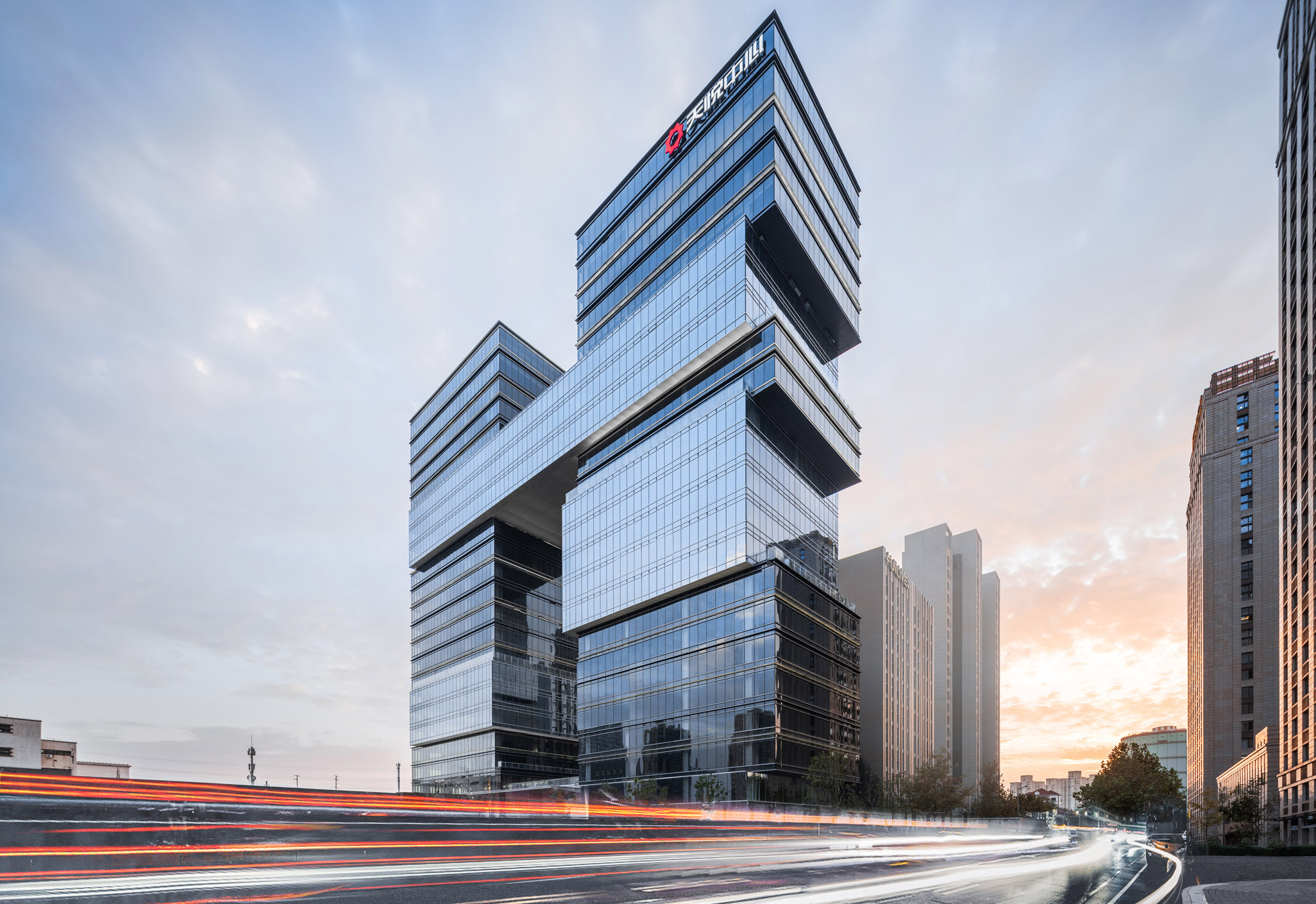 Aedas’ SCE Zhenru office towers with stacked glass volumes, cantilevered terraces and integrated sky connections in Shanghai. Street‑level dusk view of the Aedas‑designed Shanghai SCE Zhenru development, showing two office towers composed of staggered, cantilevered glass volumes. Floor‑to‑ceiling curtain wall façades define stacked blocks of varying sizes, with horizontal projections forming viewing platforms. The towers are visually linked at upper levels and front a primary urban roadway.