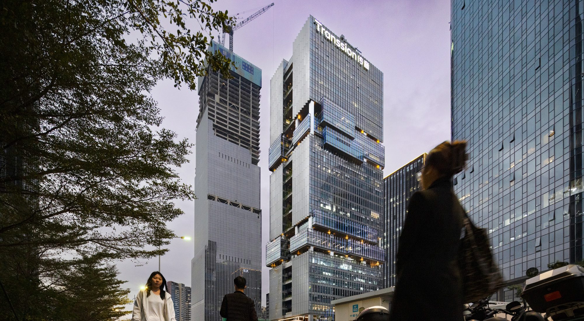 Aedas’ Shenzhen T33 Tower—office complex with glass curtain wall, illuminated sky-terrace boxes and street frontage. Street-level dusk view of Aedas’ Shenzhen T33 Tower (Liuxiandong Headquarters Base): steel-frame office tower with glass curtain wall and three irregular cantilevered ‘cloud box’ terraces. Ground plane shows pedestrian edge and scooter parking. Façade lighting and floor-to-ceiling glazing support daylighting strategy, digital workplace design and urban ecology integration on site.