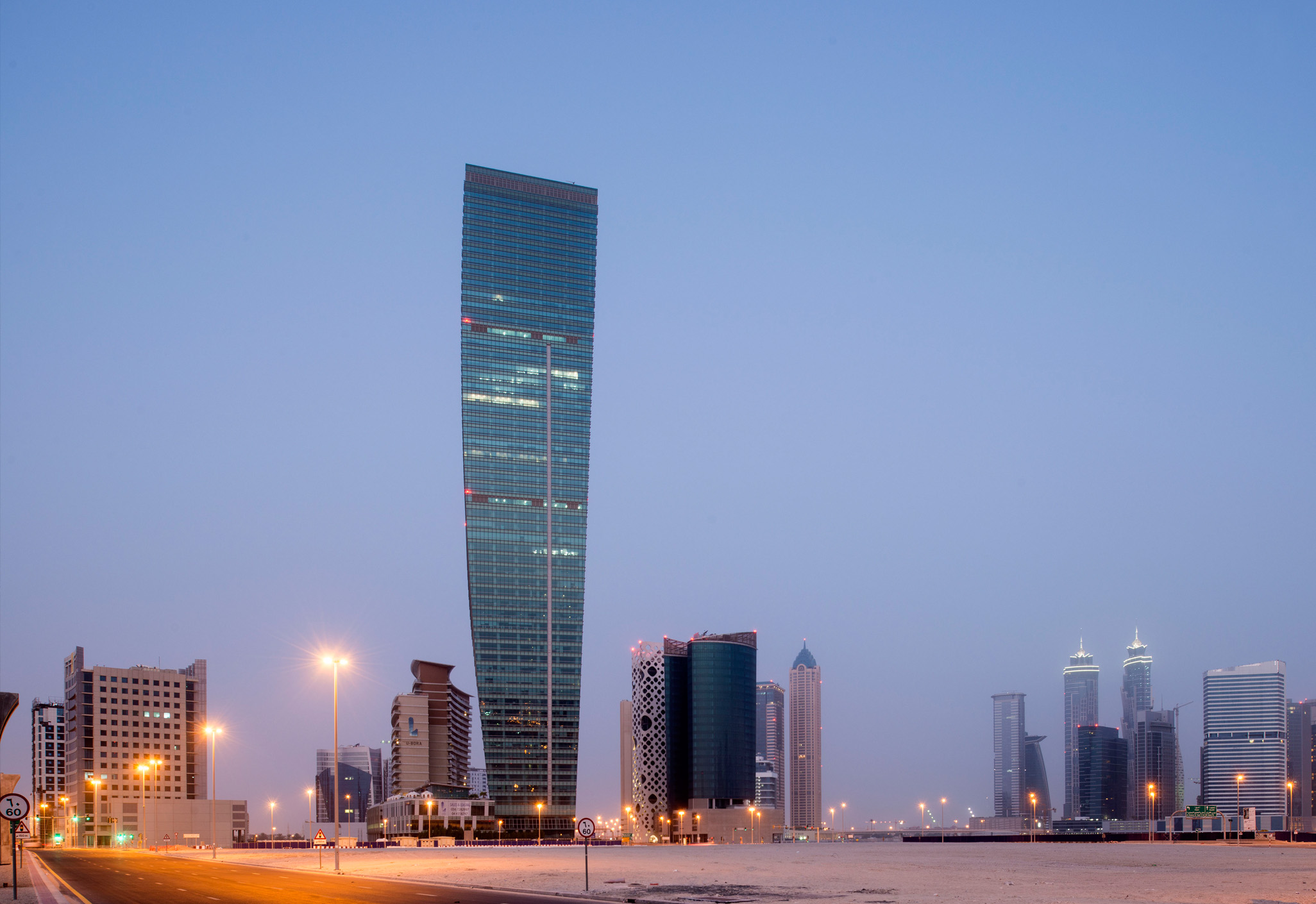 Aedas’s U‑Bora Towers in Dubai is shown as a landmark high‑rise complex dominated by a 263‑metre office tower with a gently curving, tapered form. The skyline view highlights the tower’s shallow twist, vertical glazing, and its relationship to a low‑rise podium and surrounding Business Bay towers.