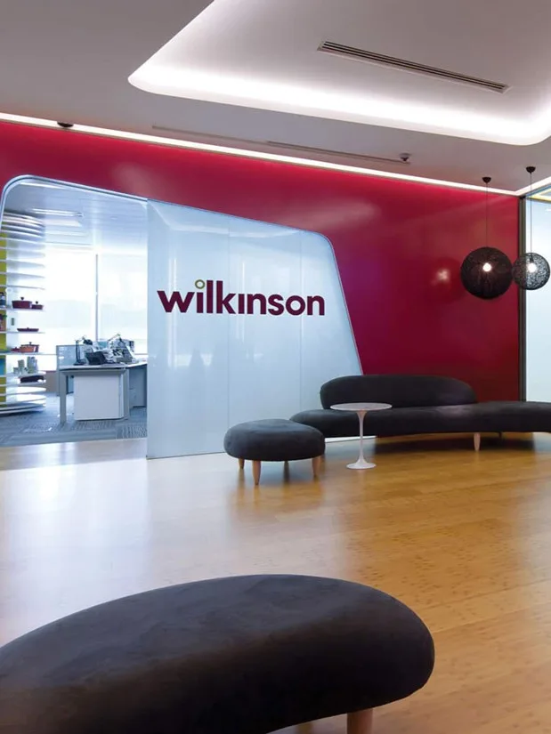 Aedas Interiors’ Wilkinson Asia Headquarters in Hong Kong is shown with a brand‑forward collaborative environment featuring a curved white innovation room, bold red feature walls, timber flooring, and glazed meeting suites. The image highlights flexible lounge seating, strong corporate branding integration, and spatial planning that prioritises informal collaboration and dynamic interaction across the workplace.