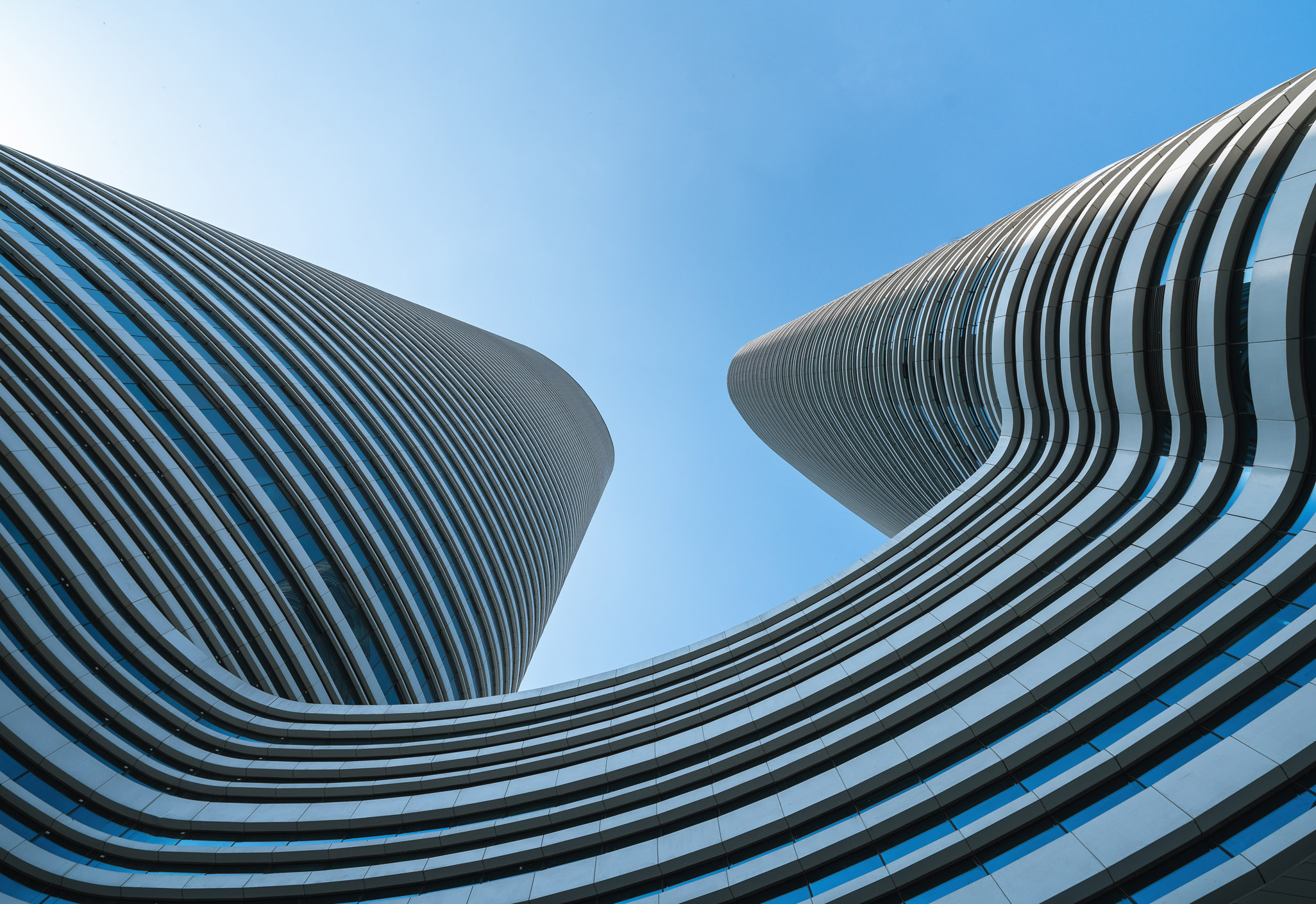 Aedas’s Xuzhou Suning Plaza is shown as a mixed‑use commercial complex with two curvilinear high‑rise towers rising from a horizontally layered retail podium. The exterior highlights continuous ring‑shaped façade bands, smooth tower profiles, and fluid massing integrated within Xuzhou’s central business district.