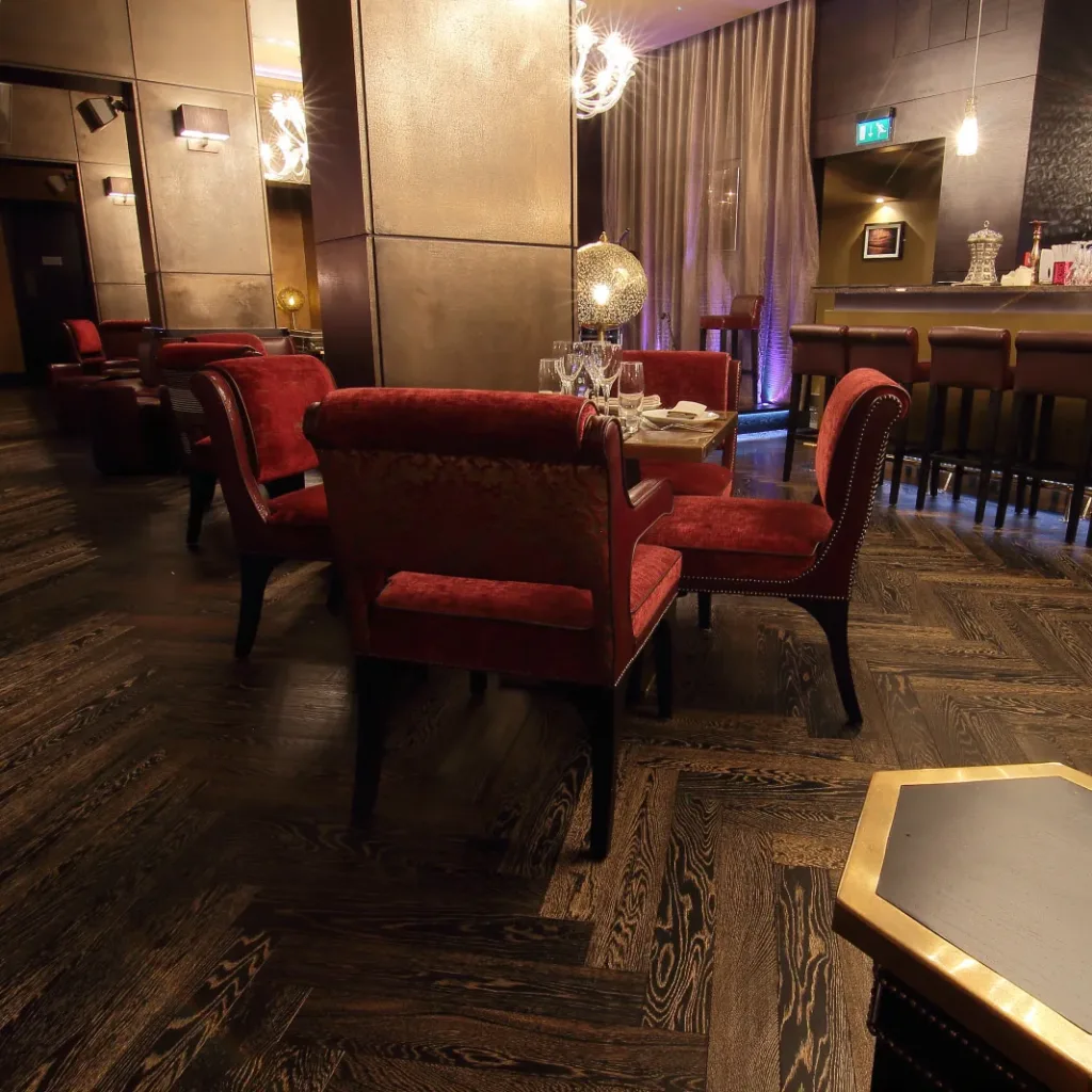 Aedas Interiors’ Mamounia Lounge in Mayfair, London is shown as an intimate hospitality interior featuring leather‑clad walls, deep red upholstered seating, dark timber flooring, and warm ambient pendant lighting. The image highlights a richly layered lounge and bar environment where tinted mirrors, sheer drapery, and low lighting create a sense of privacy, depth, and Arabic‑inspired luxury.