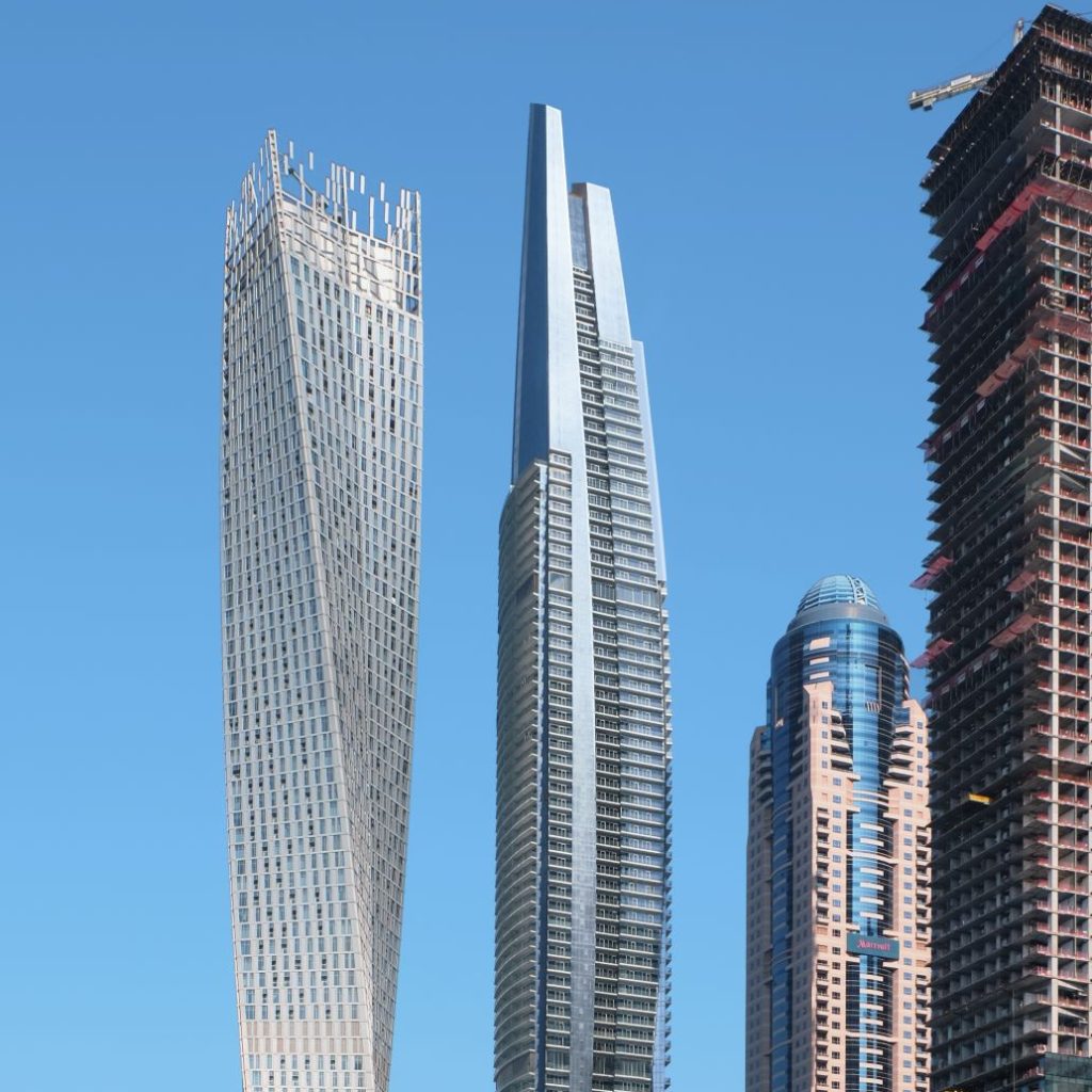 Aedas’s DAMAC Heights in Dubai is shown as a tall residential skyscraper with a vertically emphasised profile, continuous glass curtain wall, and a distinctive stepped crown. The exterior highlights high‑rise residential architecture integrated into Dubai Marina’s dense skyline, with articulated balcony bands and a tapered upper massing.