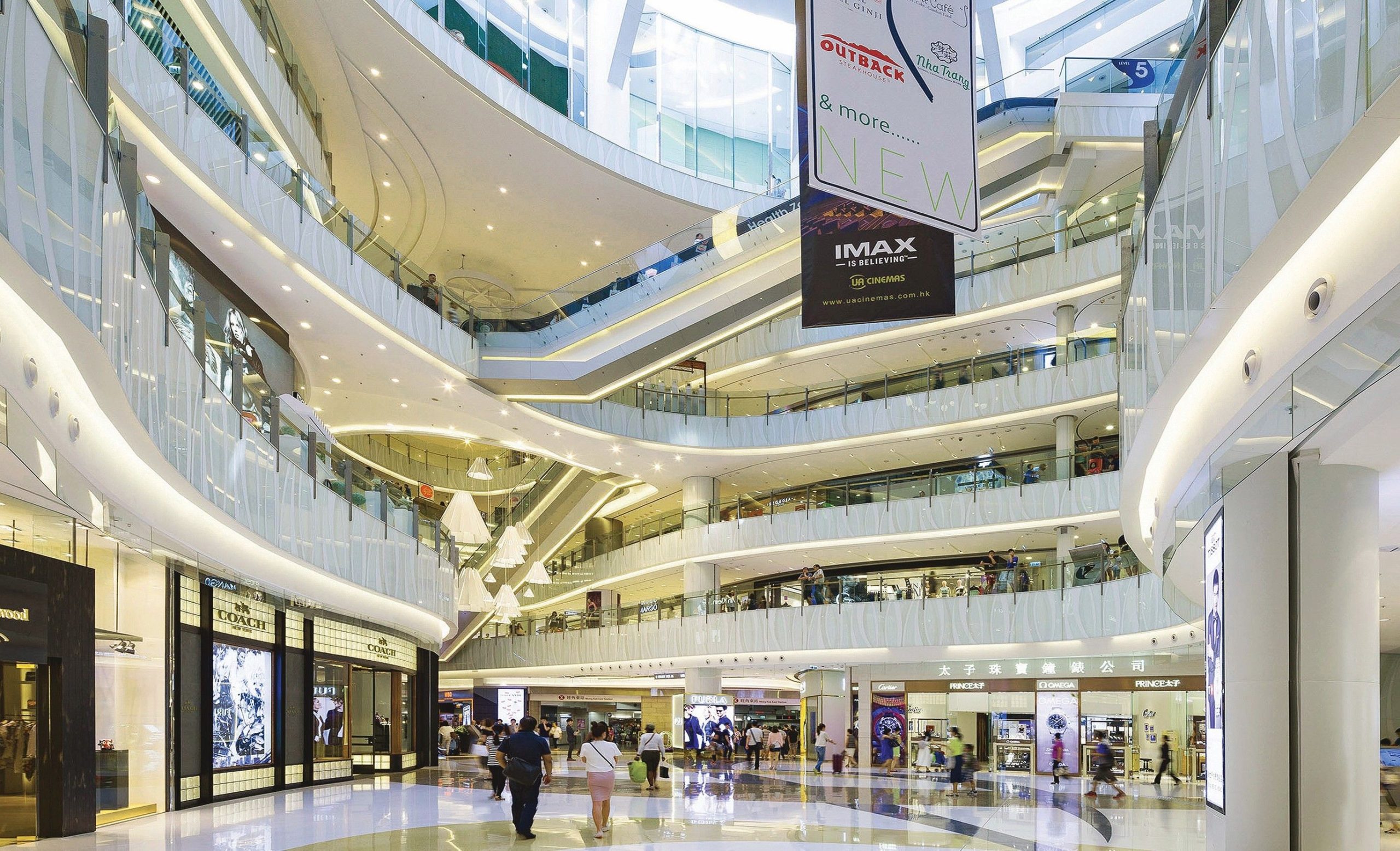 Aedas’s MOKO in Hong Kong is shown with a large multi‑storey retail atrium featuring curved slab edges, transparent balustrades, layered circulation galleries, and integrated escalators. The image highlights vertically connected mall architecture, high‑capacity public circulation, and daylight‑oriented commercial space within a dense urban context.
